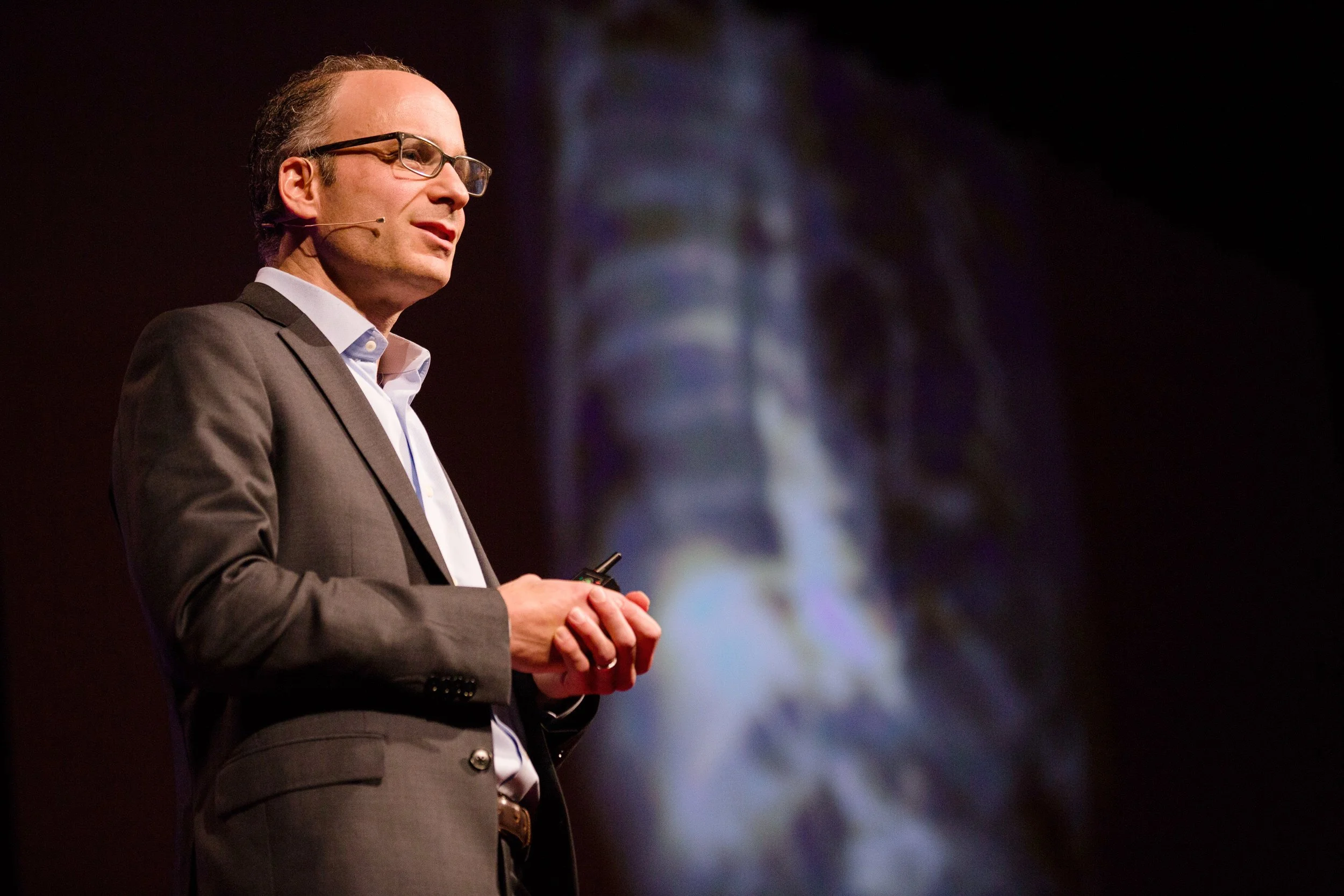A man in a gray suit and glasses giving a TEDx presentation on stage, with a projection of a human spine in the background, an an event in Boston, Massachusetts.