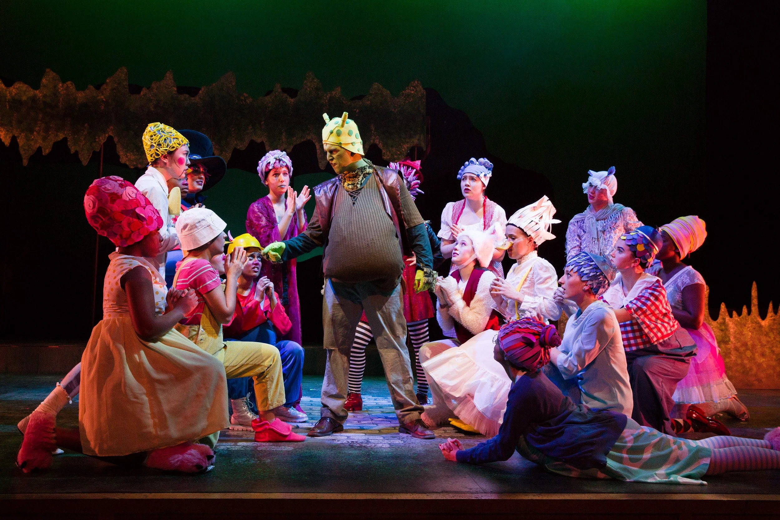 A stage scene from Shrek musical with high school actors in colorful costumes and vibrant makeup, portraying a story in an imaginative setting.