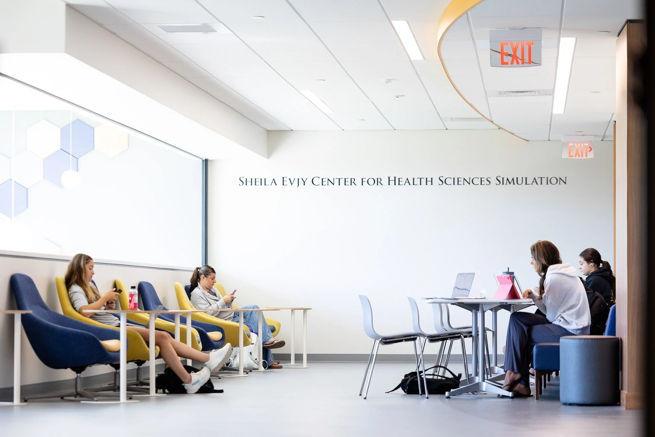 College campus lounge with four young women seated, in a modern academic building interior at Sheila Evjy Center for Health Sciences Simulation.
