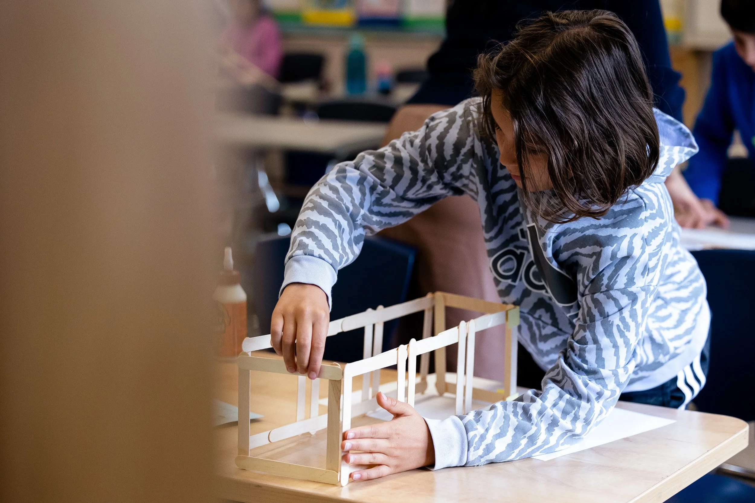 A student is working on a small model of a building made of wooden sticks on a table in a classroom.