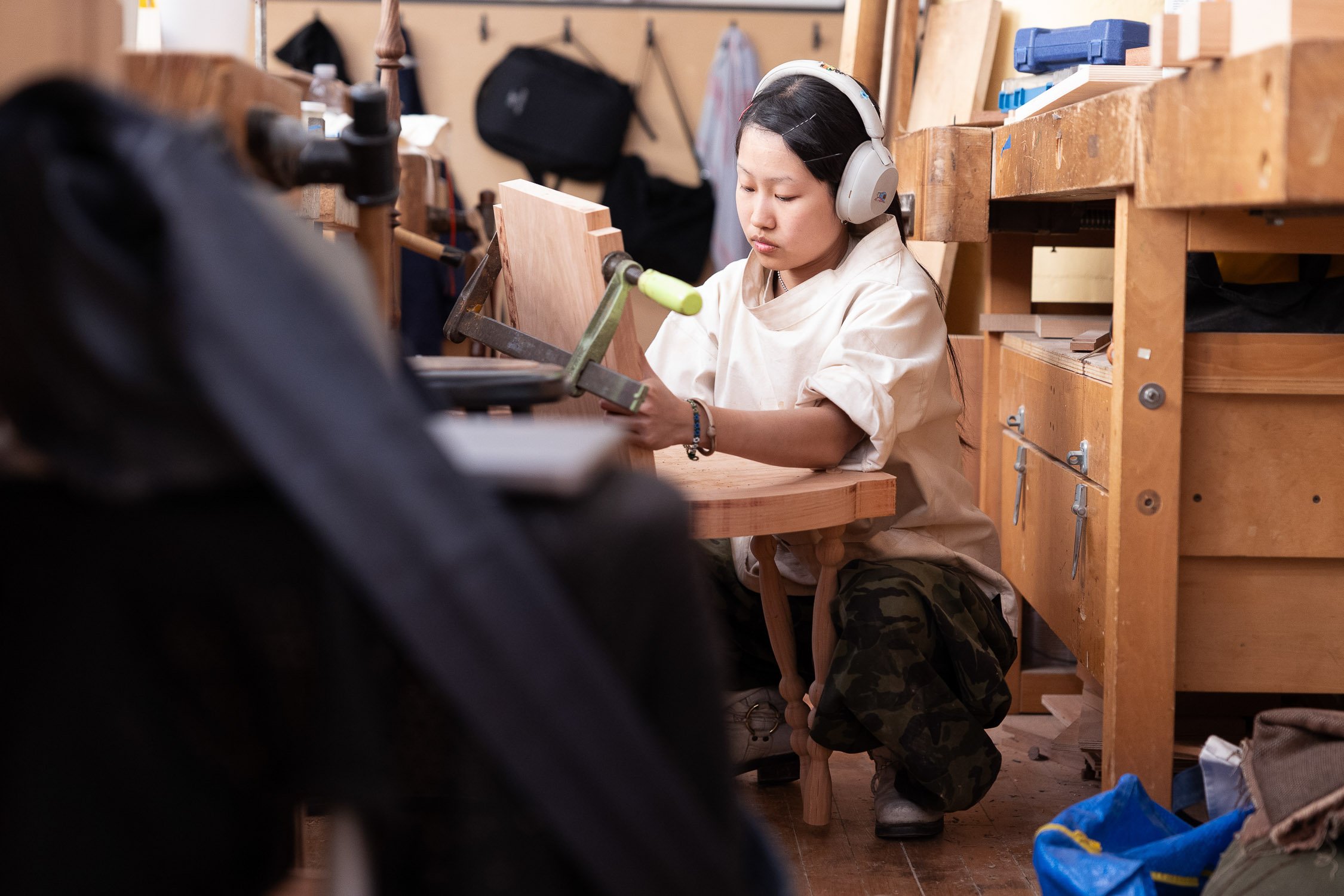 A young woman working in a student woodworking shop, wearing headphones, a beige shirt, and camouflage pants, using a saw on a piece of wood at an art school in Providece, Rhode Island.