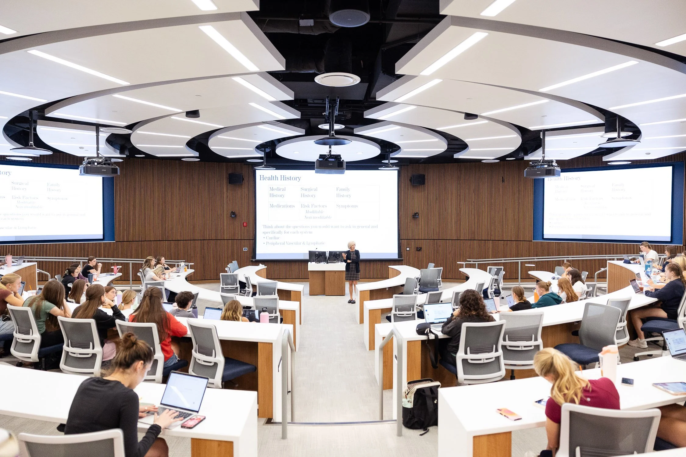 A university lecture hall filled with students seated at curved desks, listening to professor standing at the front.