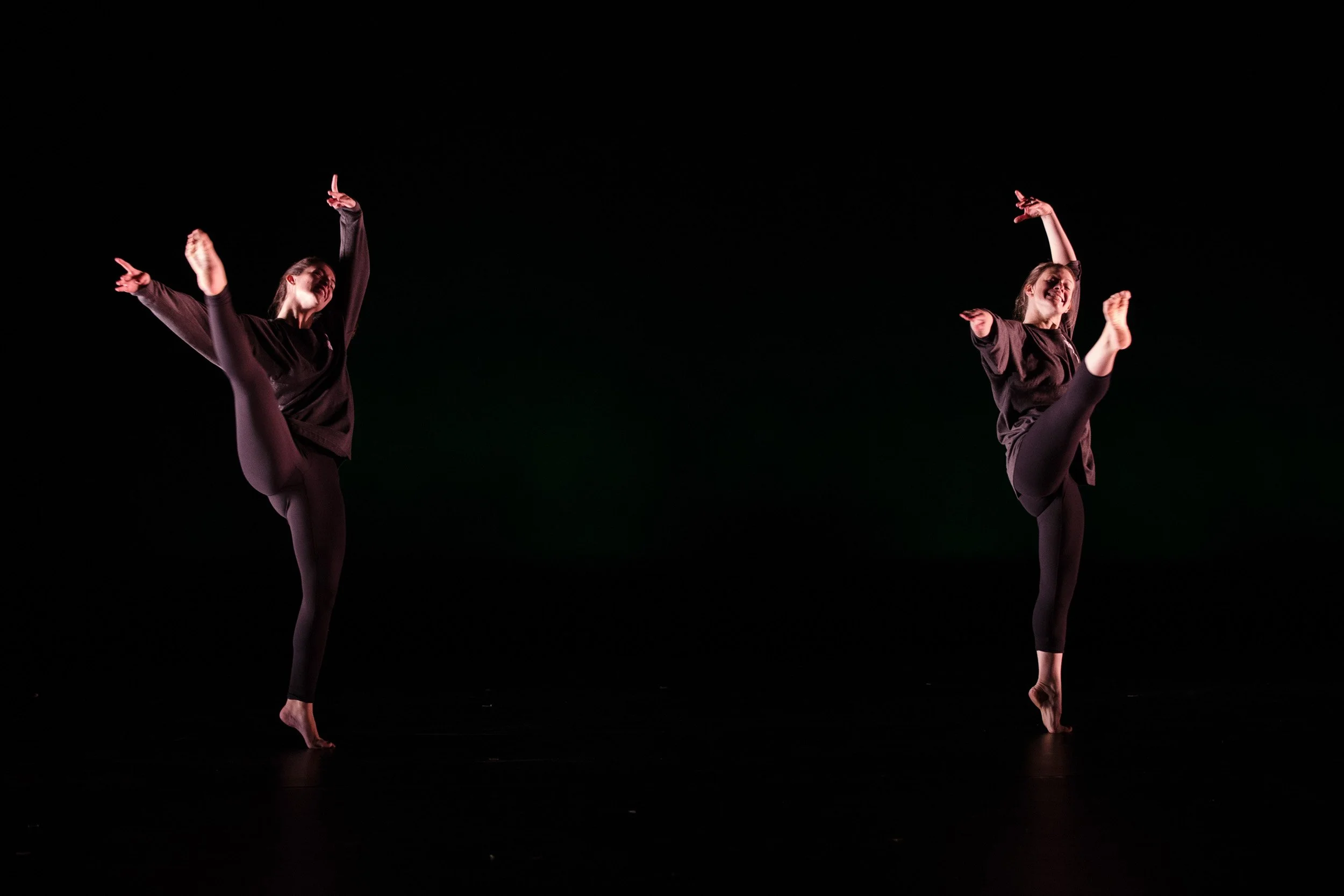 Two female dancers in black outfits performing contemporary dance on a dark stage, one with arms raised and extended, the other with arms bent and upward.