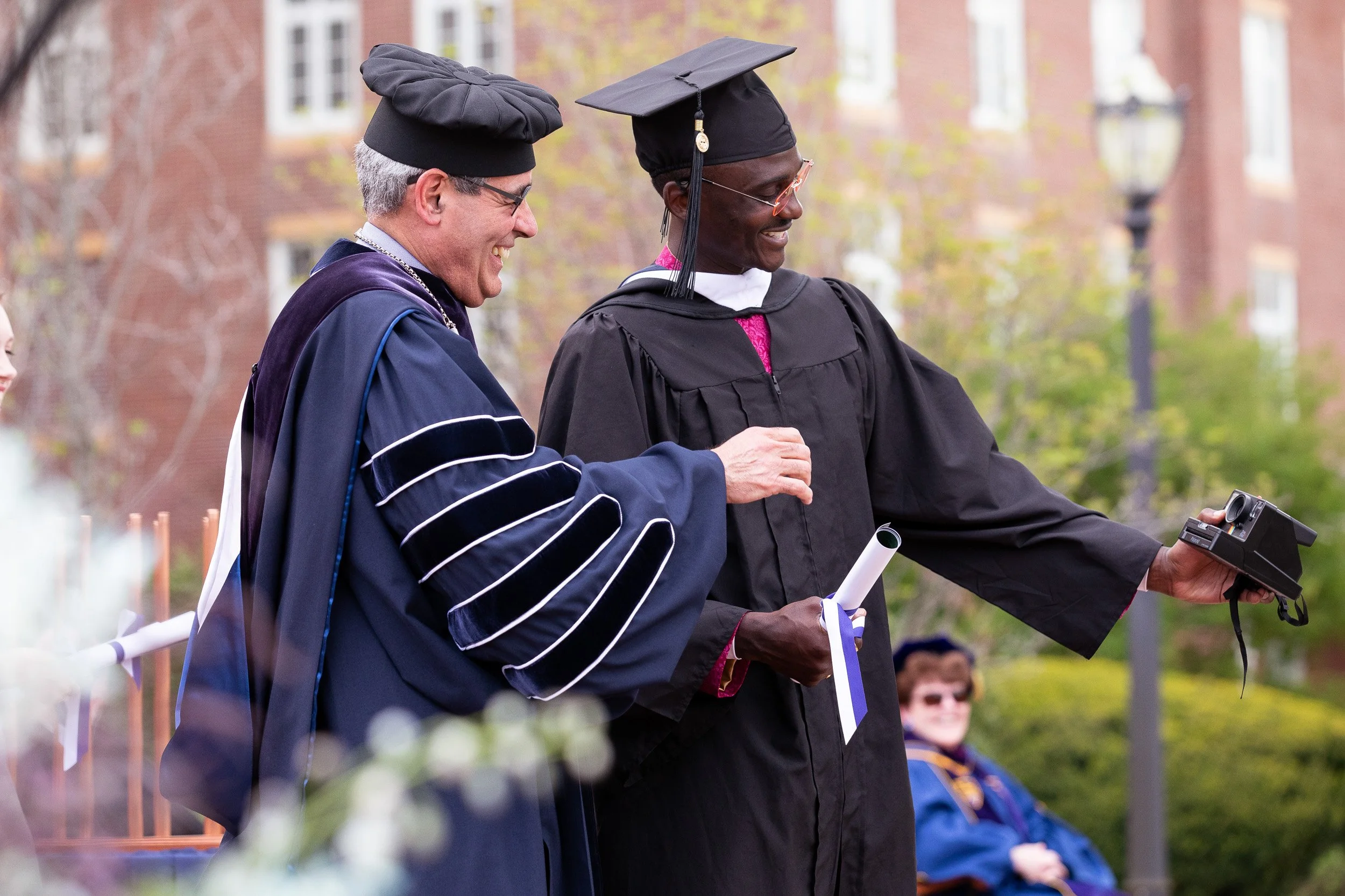 Two men at a graduation ceremony, one in academic regalia and the other in cap and gown, smiling and holding a diploma and a camera
