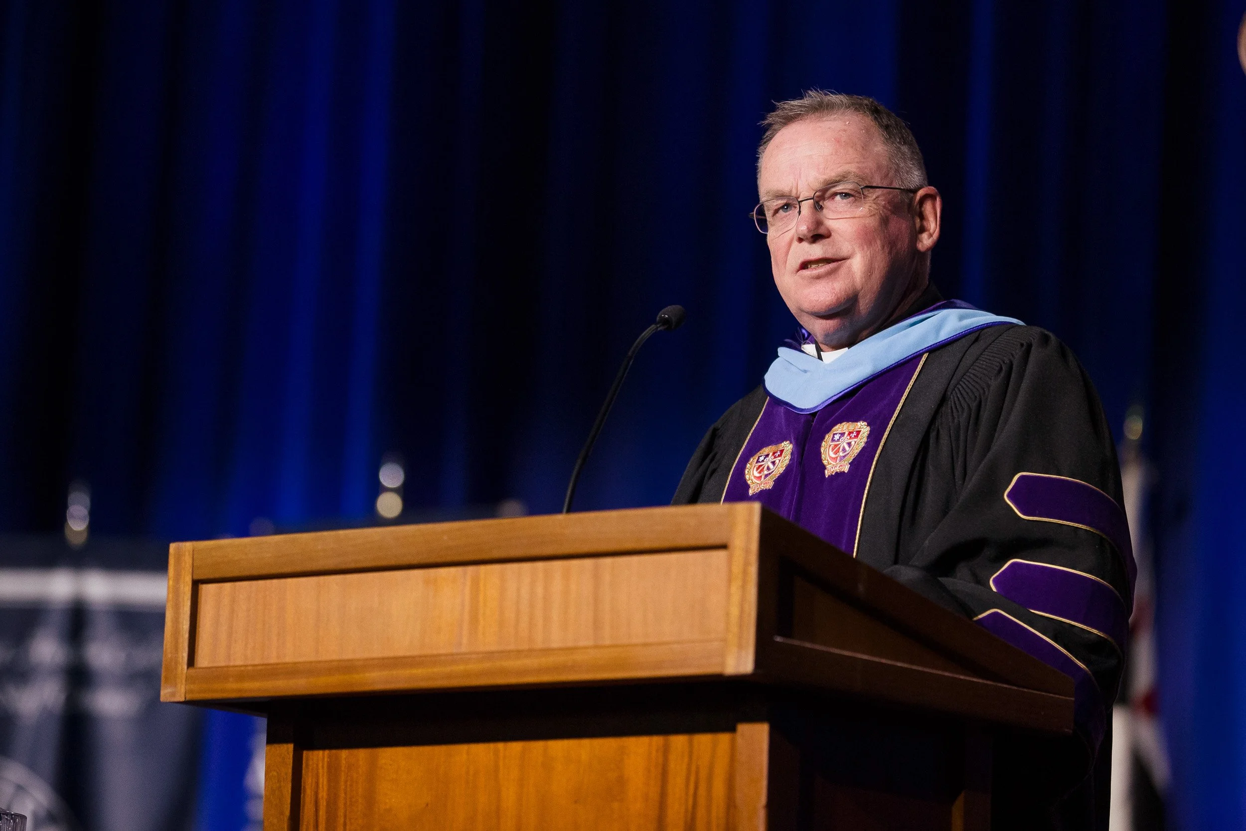 A man in academic regalia giving a speech at a wooden podium on a stage during college president inauguration.