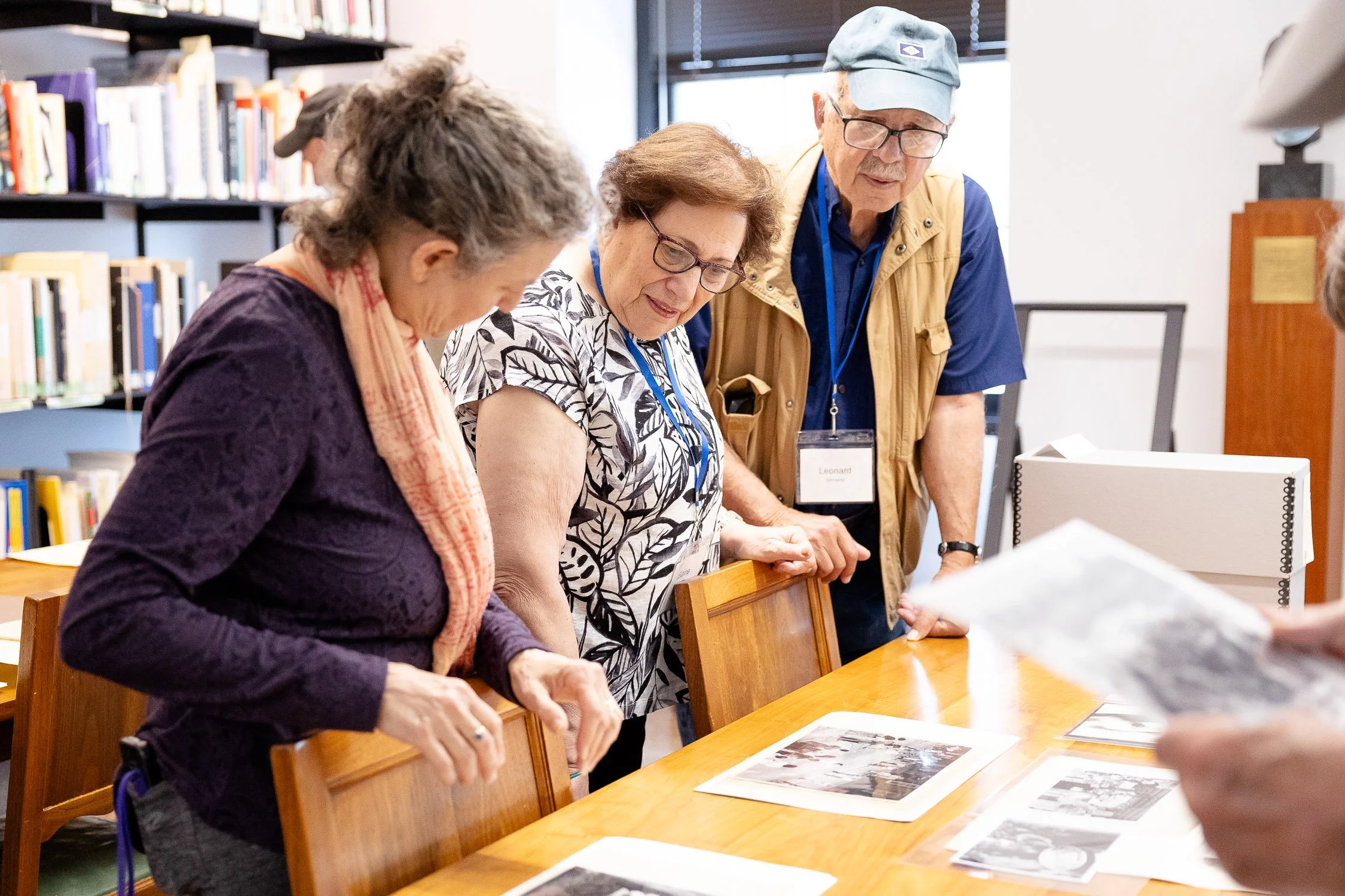 Three elderly people standing around a wooden table, looking at photographs laid on the table, at RISD reunion.