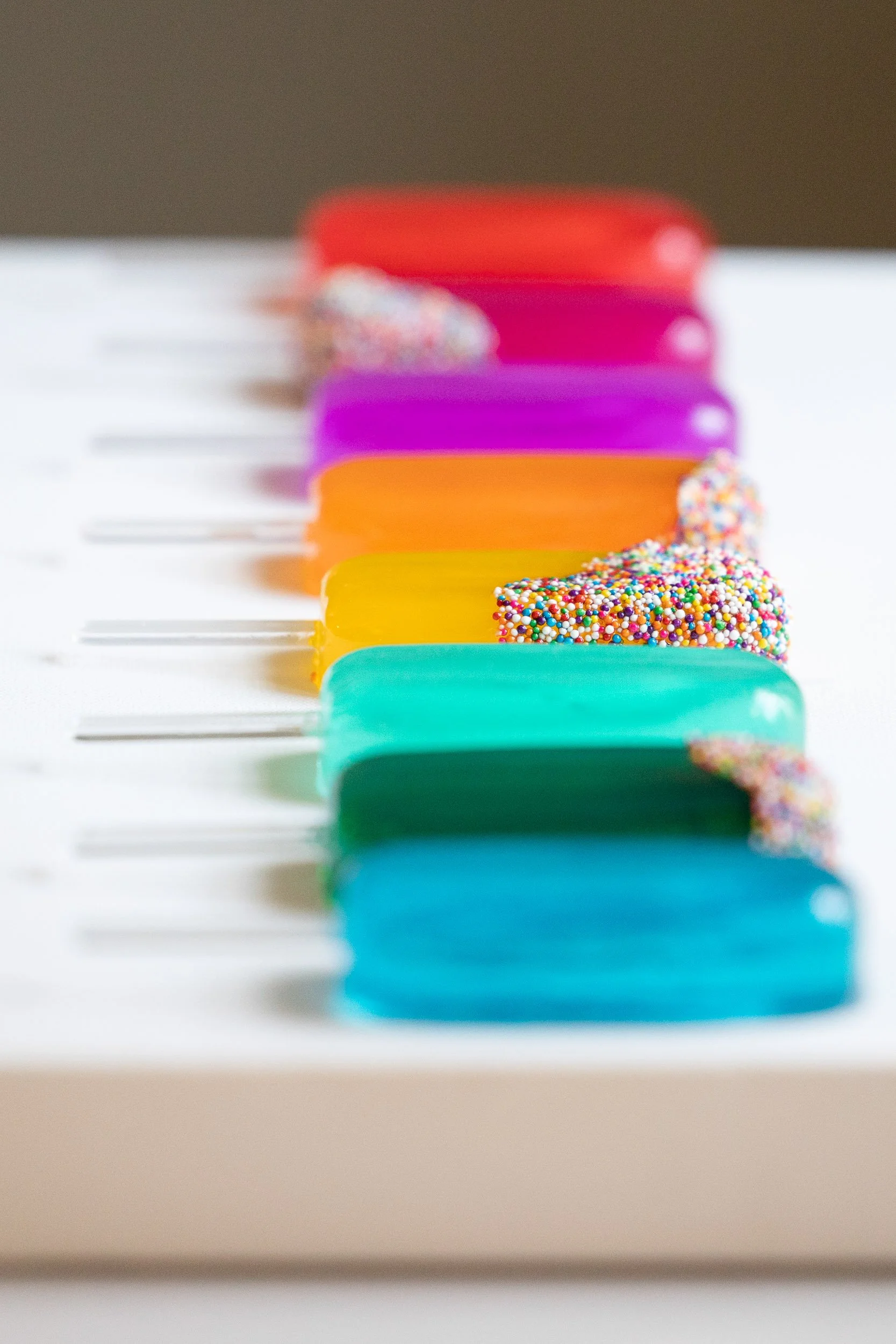 Colorful resin popsicles lined up on a white surface, with some topped with multicolored sprinkles.