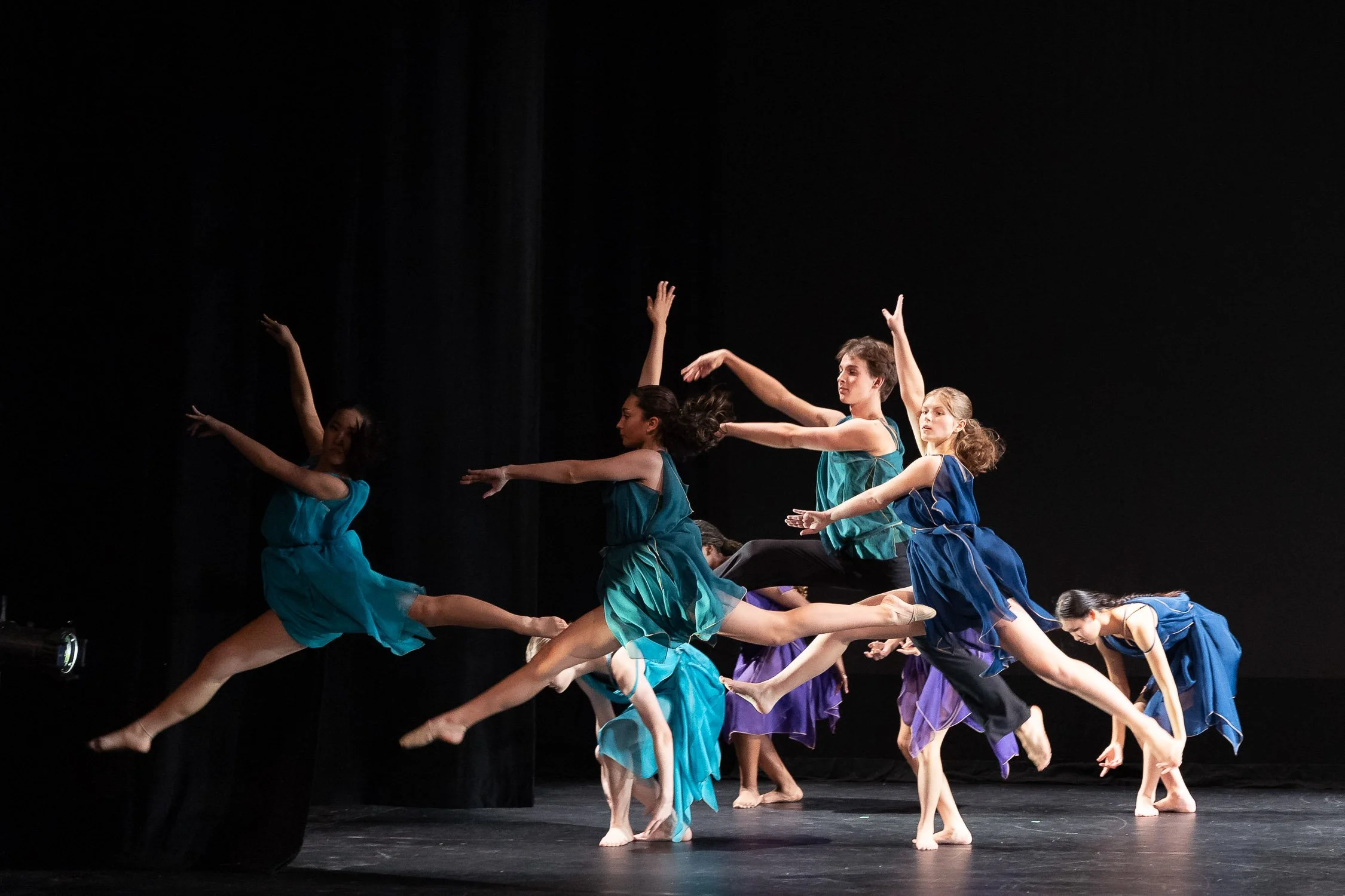 A group of high school ballet dancers performing on stage wearing blue and purple costumes, leaping and balancing with graceful poses at a dance academy in Massachusetts.