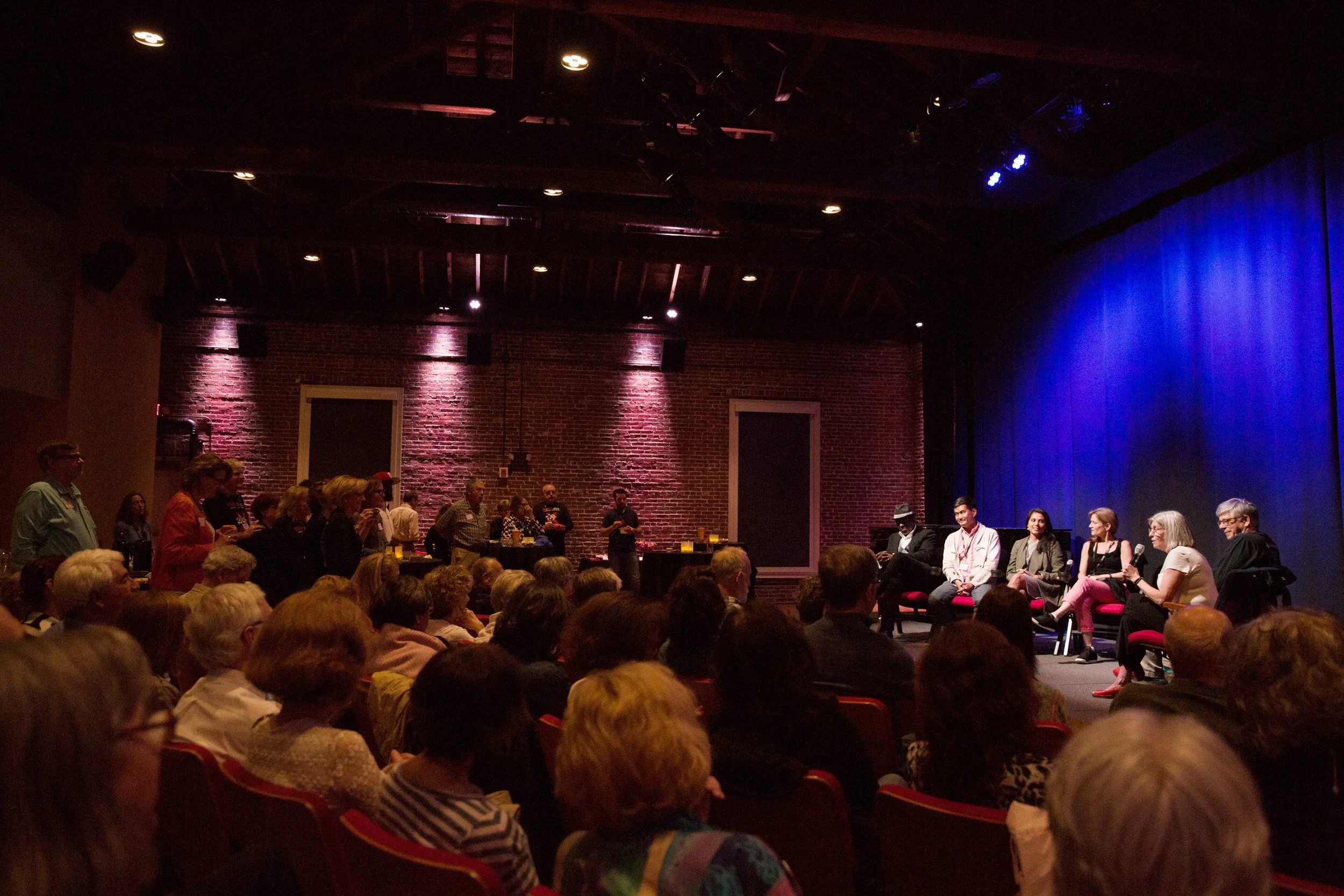 A panel discussion taking place on stage in front of an audience in a dimly lit theater with blue lighting and brick walls.