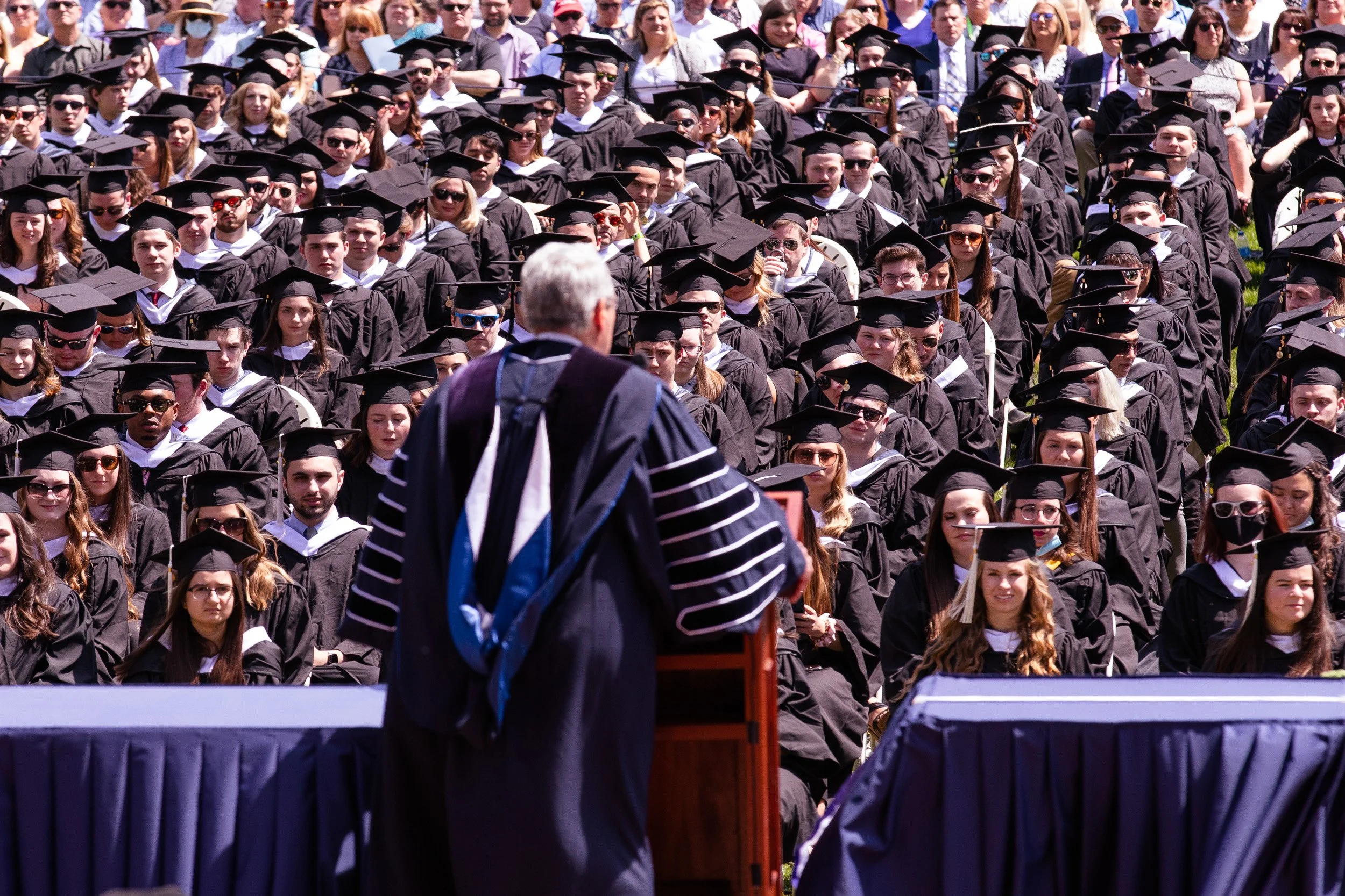 College graduation ceremony with students in caps and gowns seated outdoors, facing a speaker at a podium.