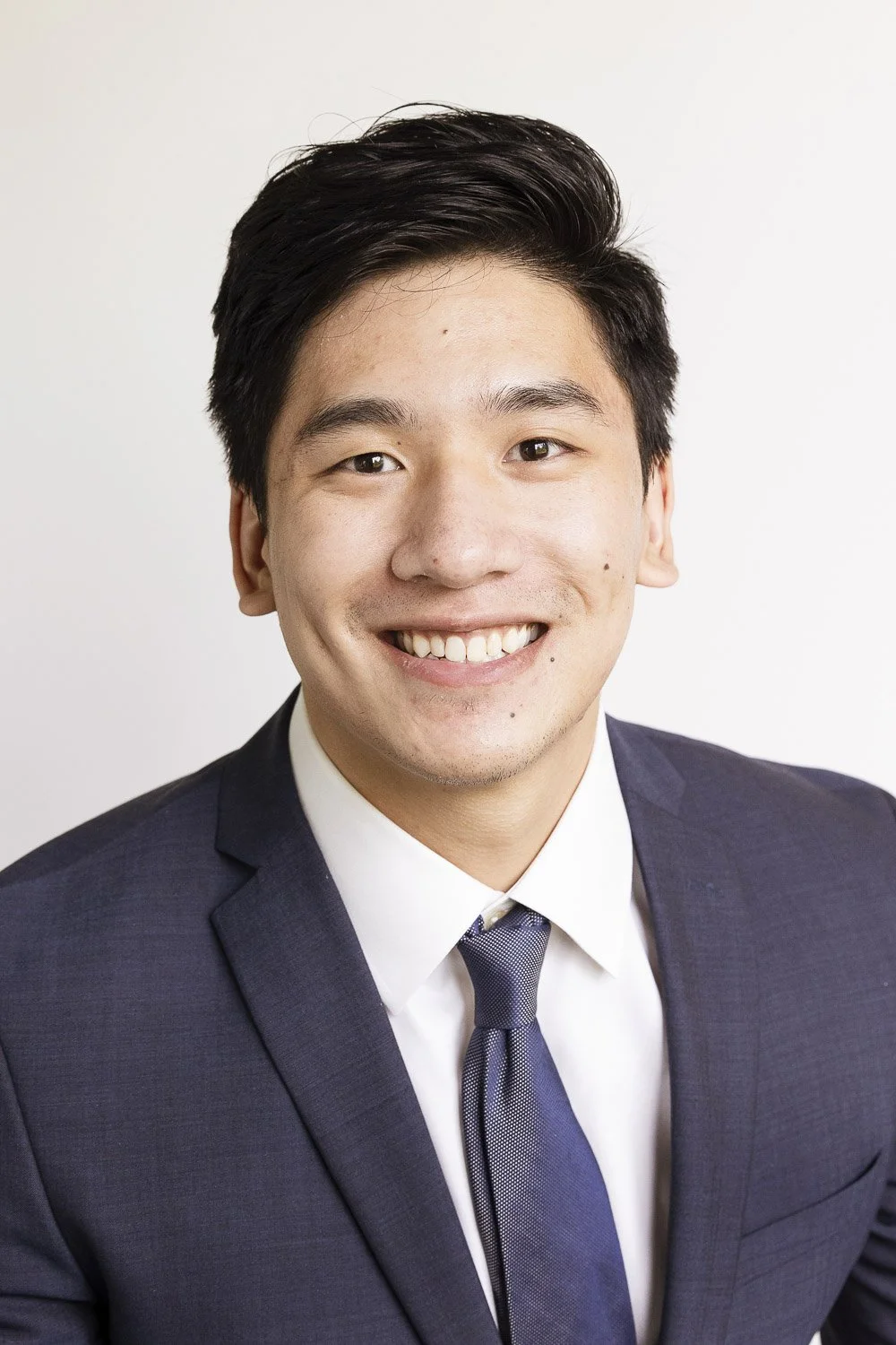 Headshot of a young man in a navy suit, white shirt, and gray tie, smiling against a plain white background.