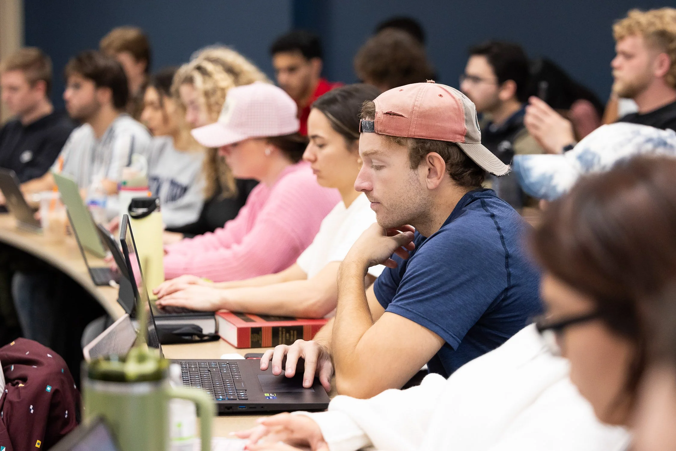 Students in a lecture hall sitting at desks with laptops, some with notebooks, appearing attentive and engaged during class.