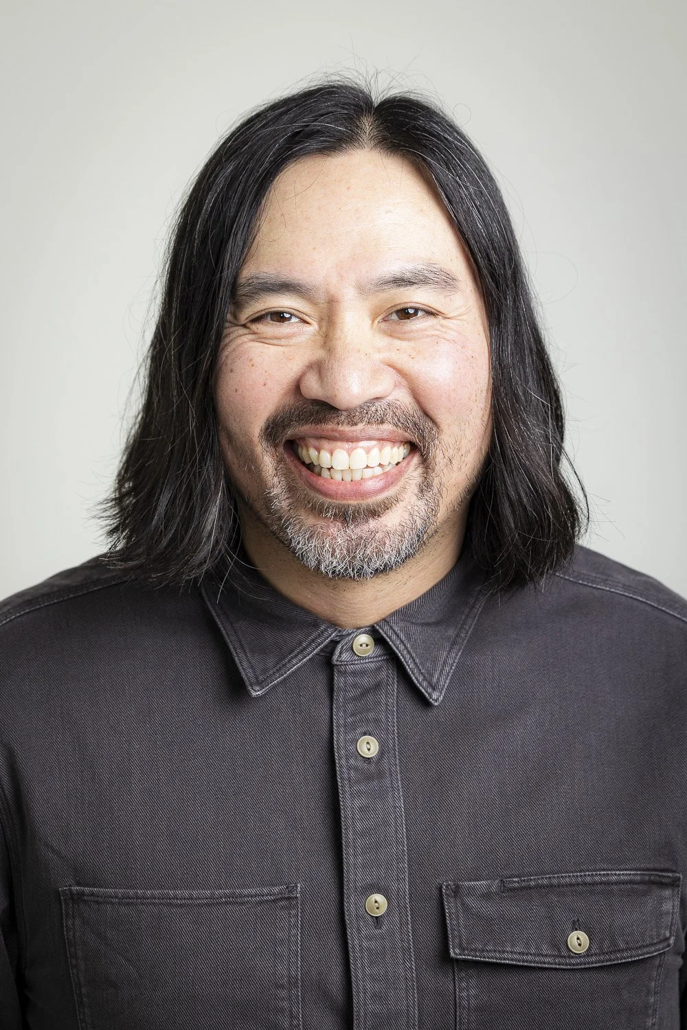 A man with long black hair, wearing a dark gray button-up shirt, smiling widely, against a plain light-colored background.
