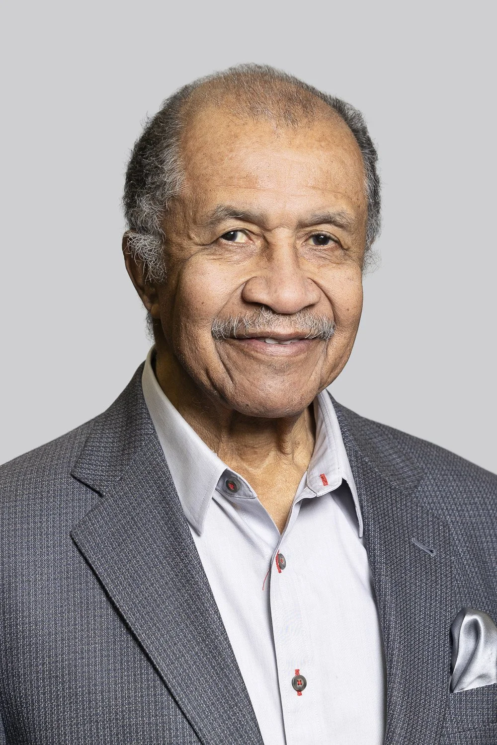 Portrait of a man with gray hair and a mustache, wearing a gray suit jacket and a light gray shirt, smiling at the camera against a plain gray background.