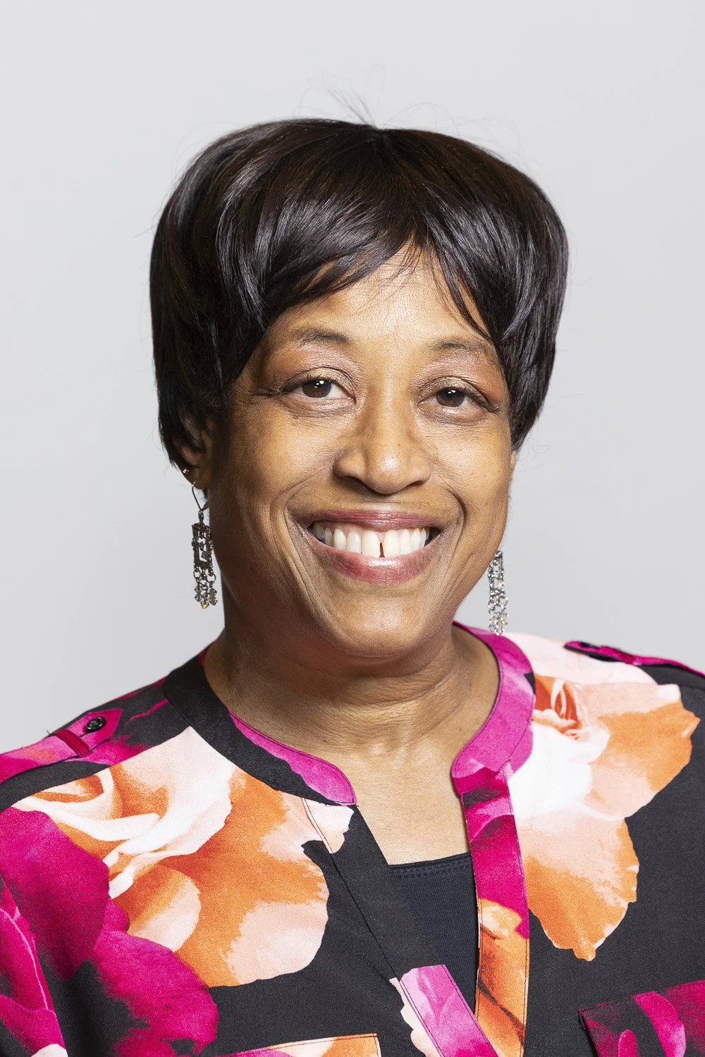 A smiling independent school alumni with short black hair, wearing earrings and a colorful floral blouse against a plain background.