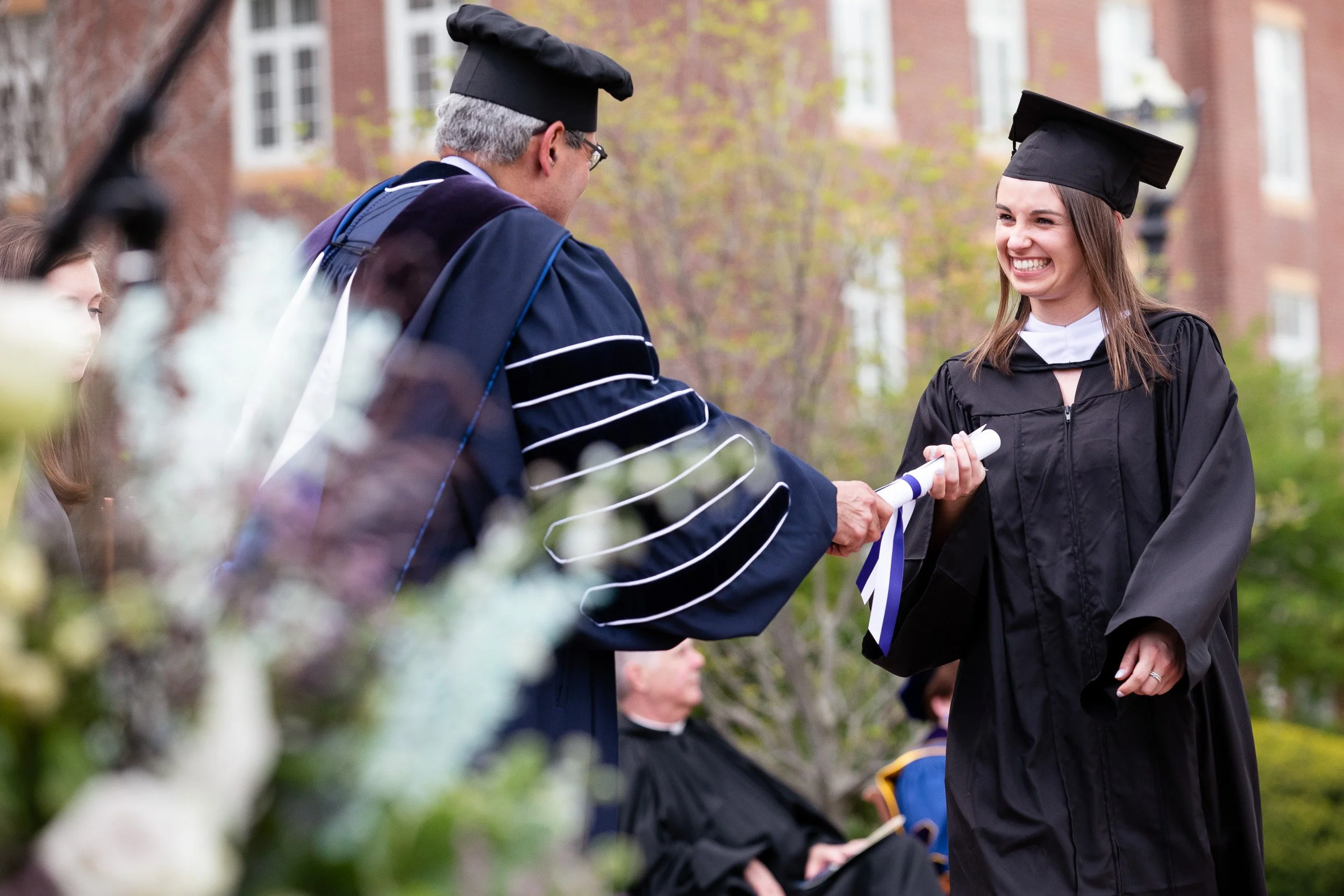 A young woman in a black graduation gown smiling as she receives her diploma at an outdoor graduation ceremony on a college campus.