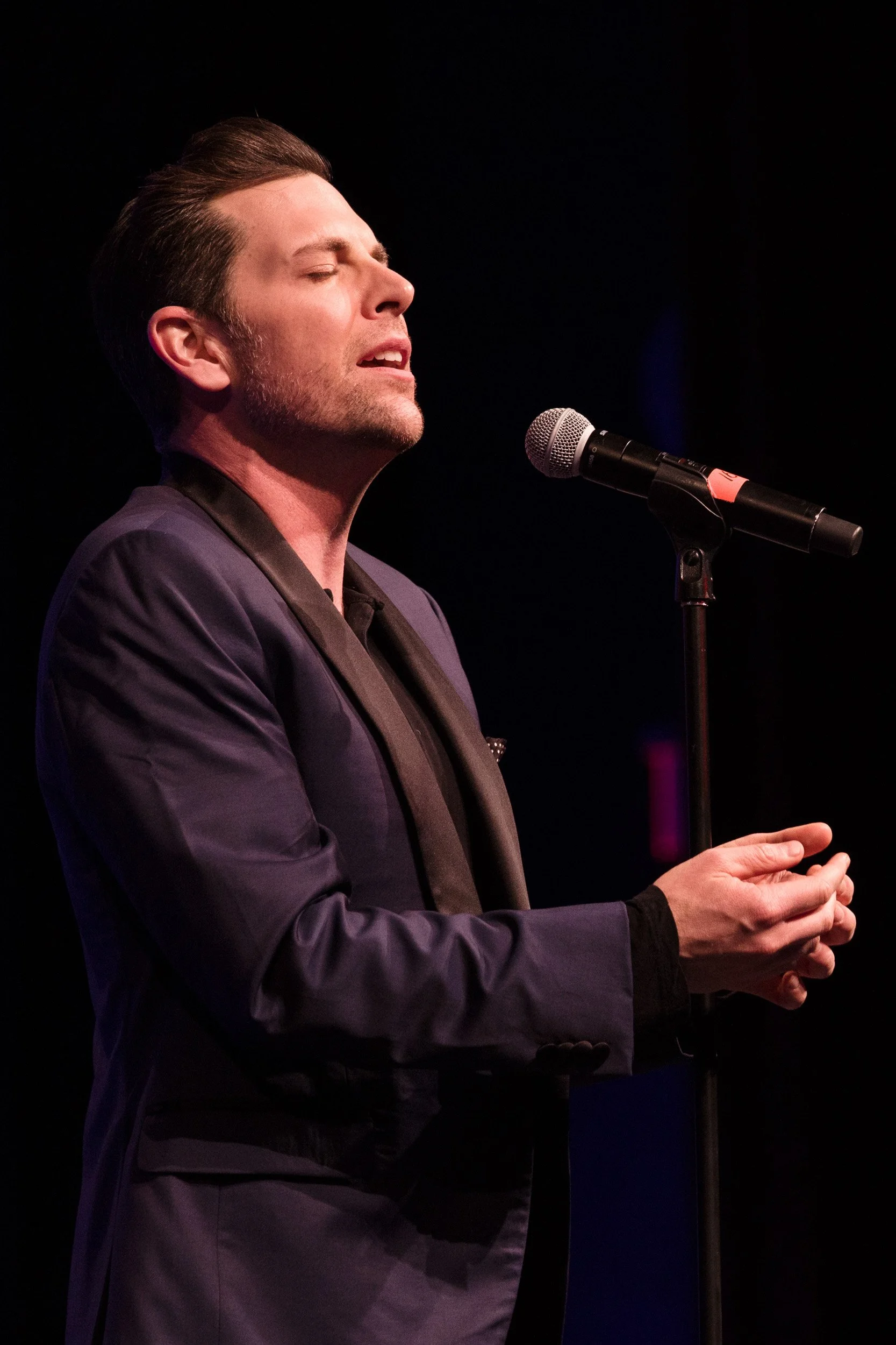 Chris Mann singing passionately on stage with eyes closed, dressed in a dark suit, holding his hands together near a microphone.