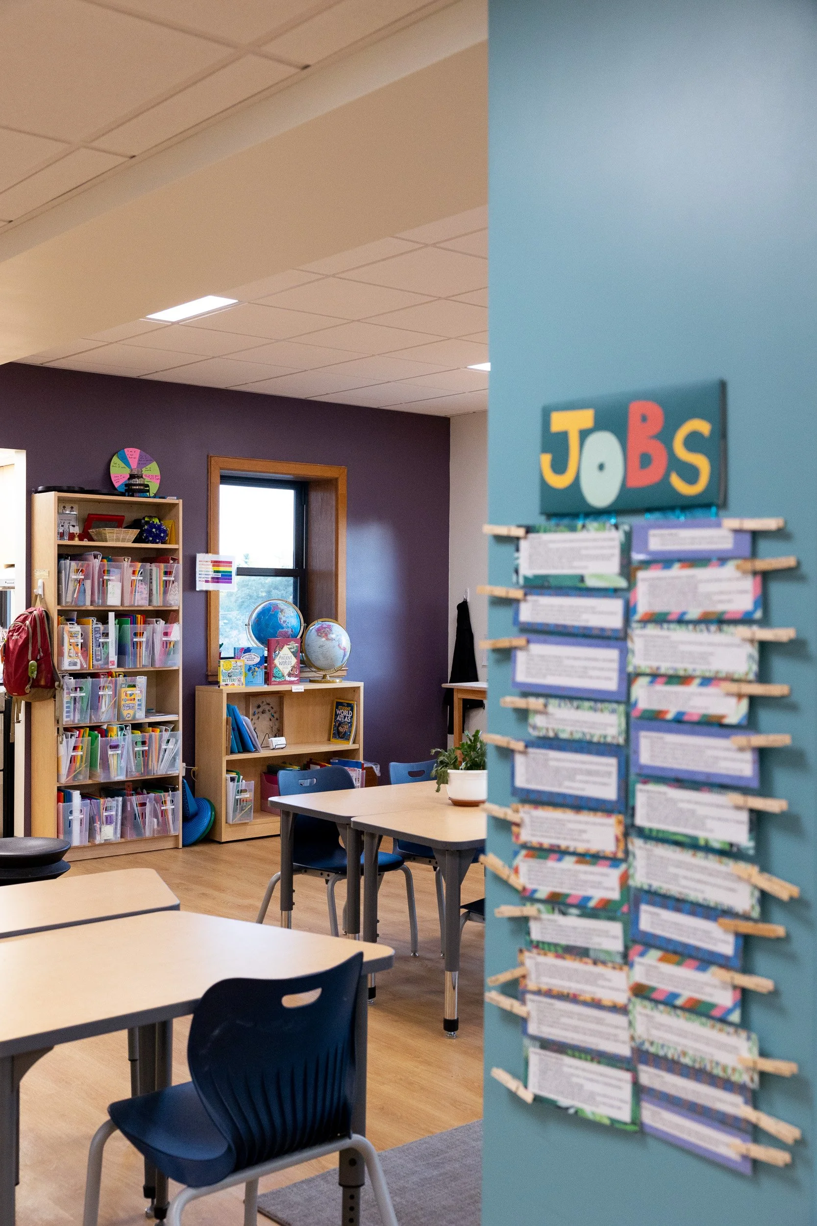 Montessori classroom with educational materials, bookshelves, globes, and a sign labeled 'JOBS' on a blue wall.