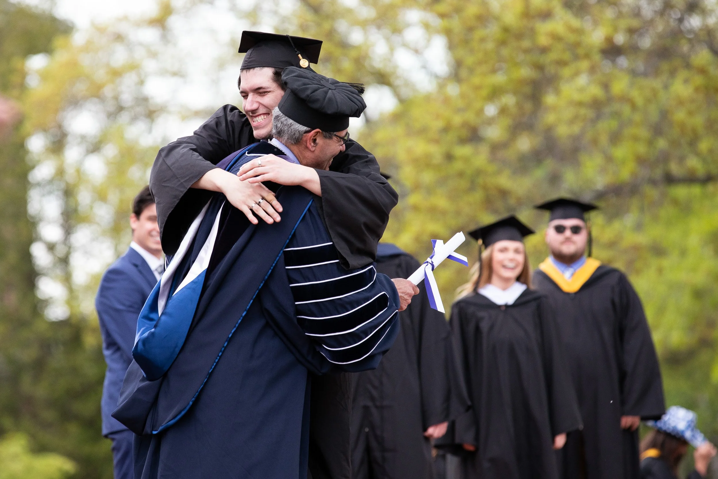 A graduate in cap and gown hugs college president during a graduation ceremony outdoors.