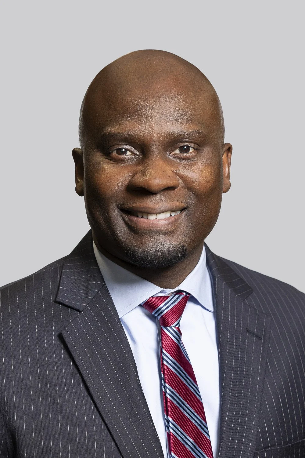 Professional headshot of a Boston law school trustee man in a dark pinstripe suit, white shirt, and striped tie against a light gray background.