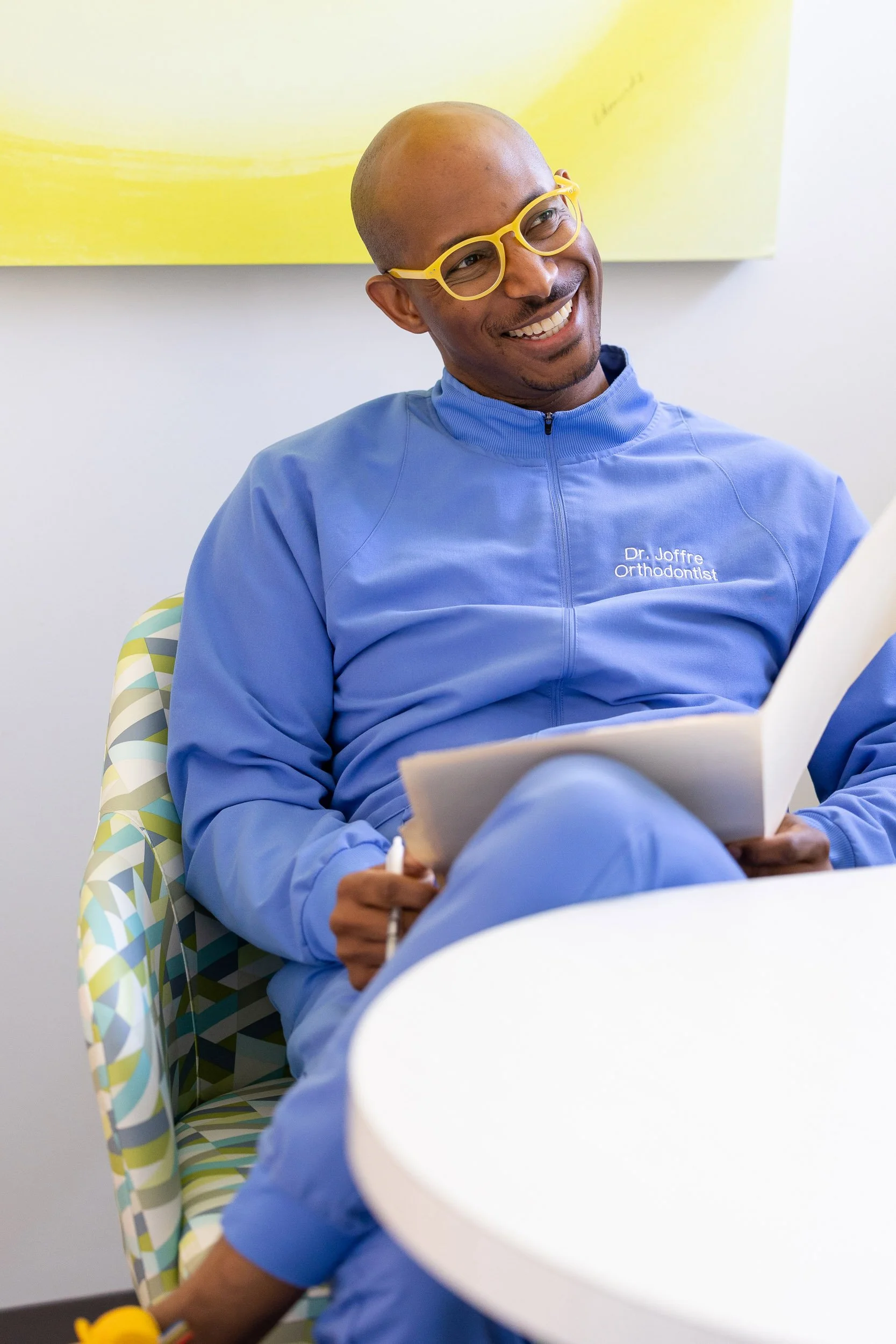 A smiling orthodontist with glasses wearing a blue jacket, sitting in a colorful patterned chair, holding a folder, in a bright room with white and yellow decor.