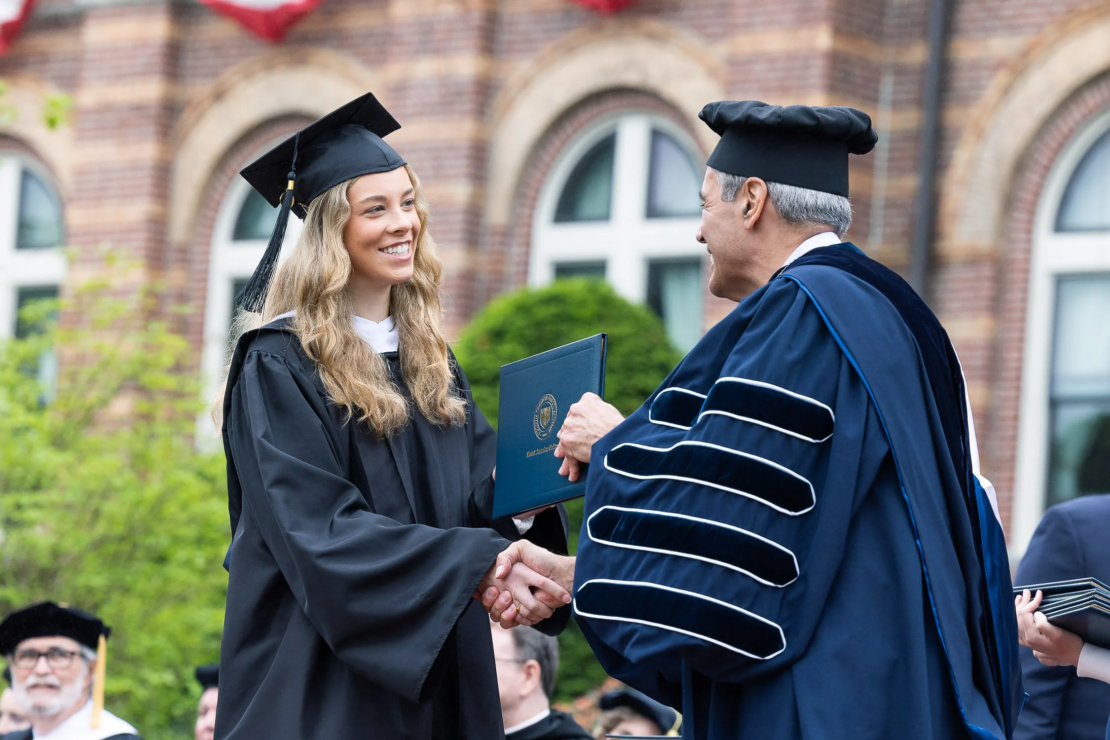 Young woman in graduation gown and cap shaking hands with college president during a graduation ceremony outside.