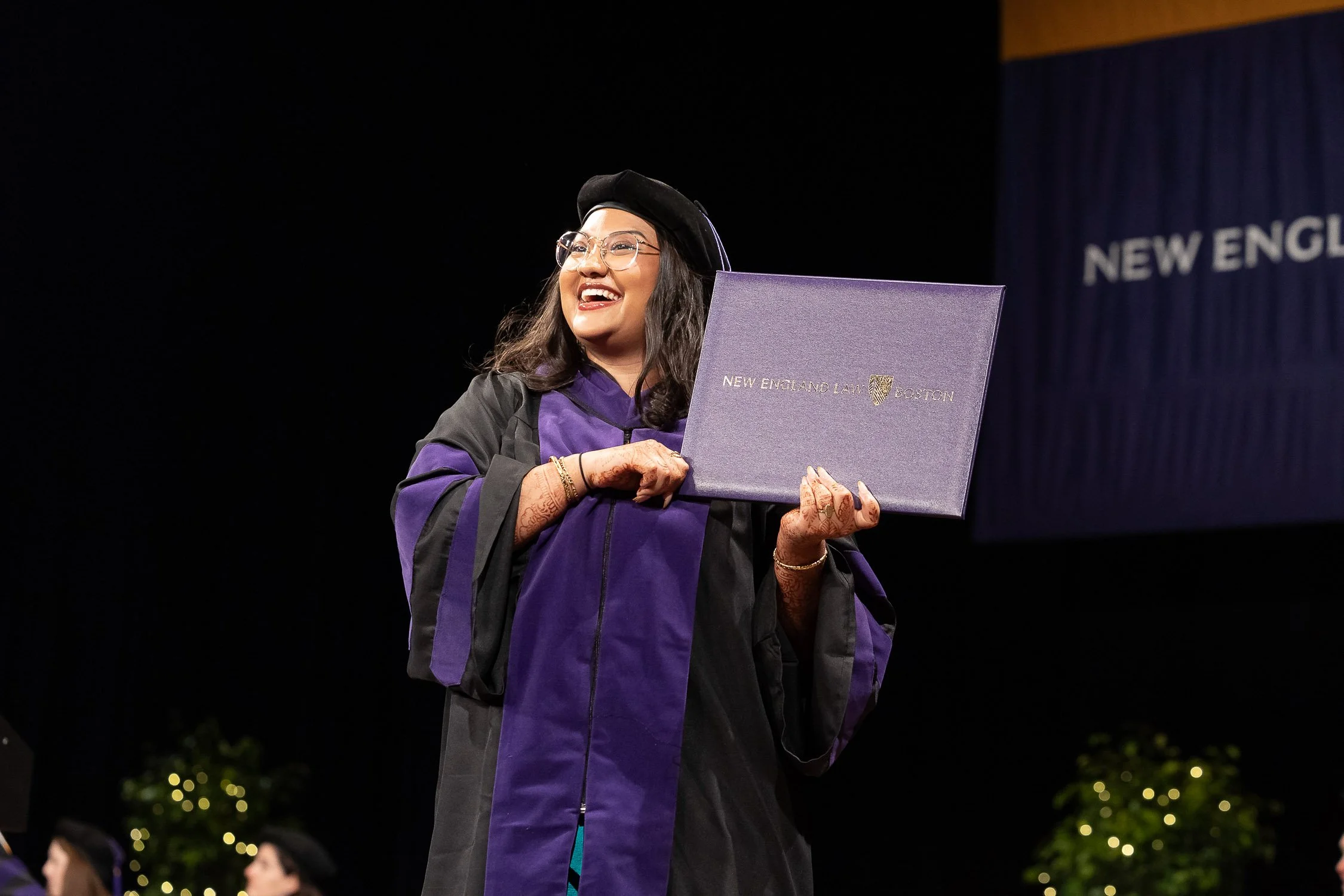 A woman in graduation regalia holding a diploma and smiling at a graduation ceremony in Boston.