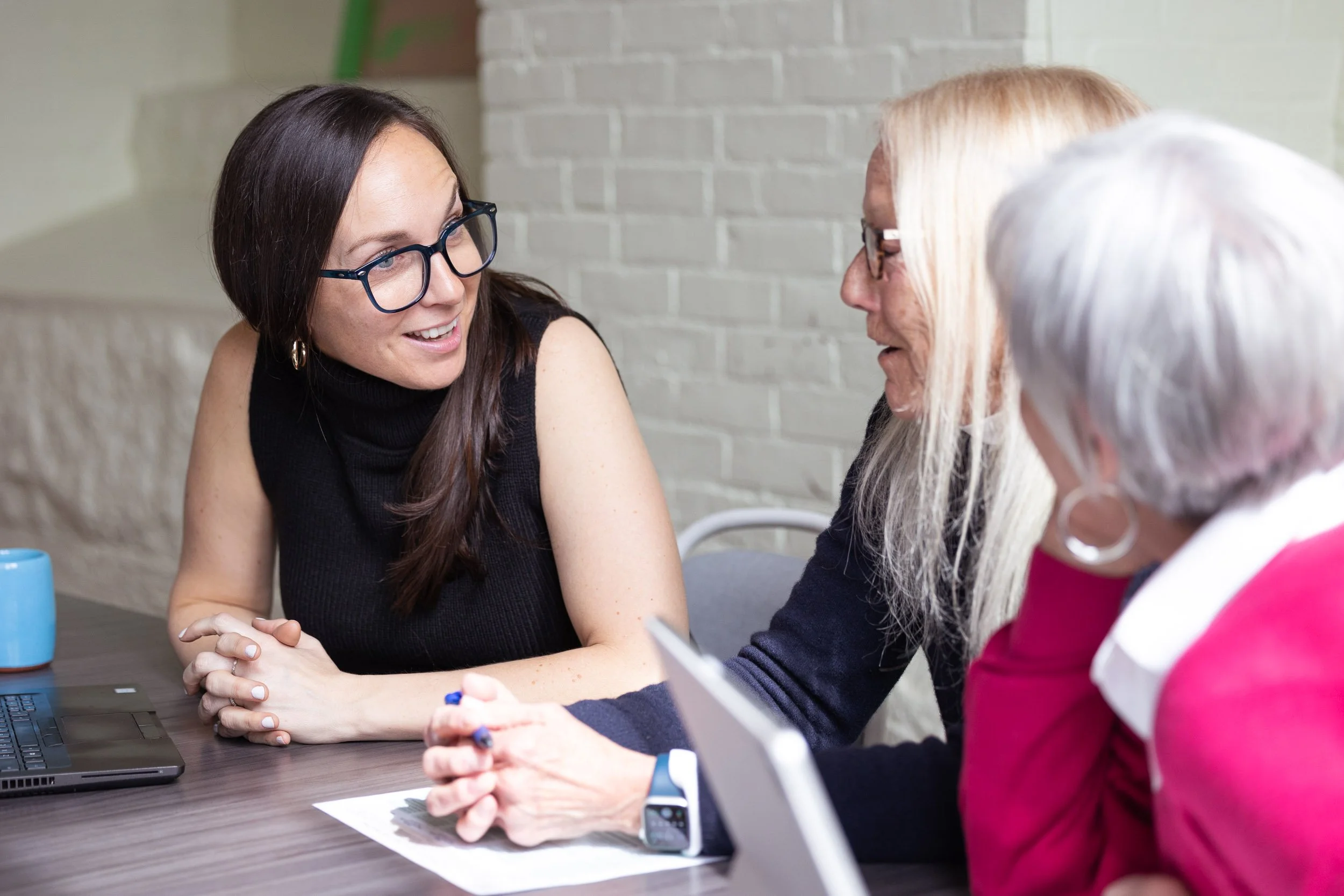 Three women having a conversation at a table, with a laptop and documents, in an office setting.