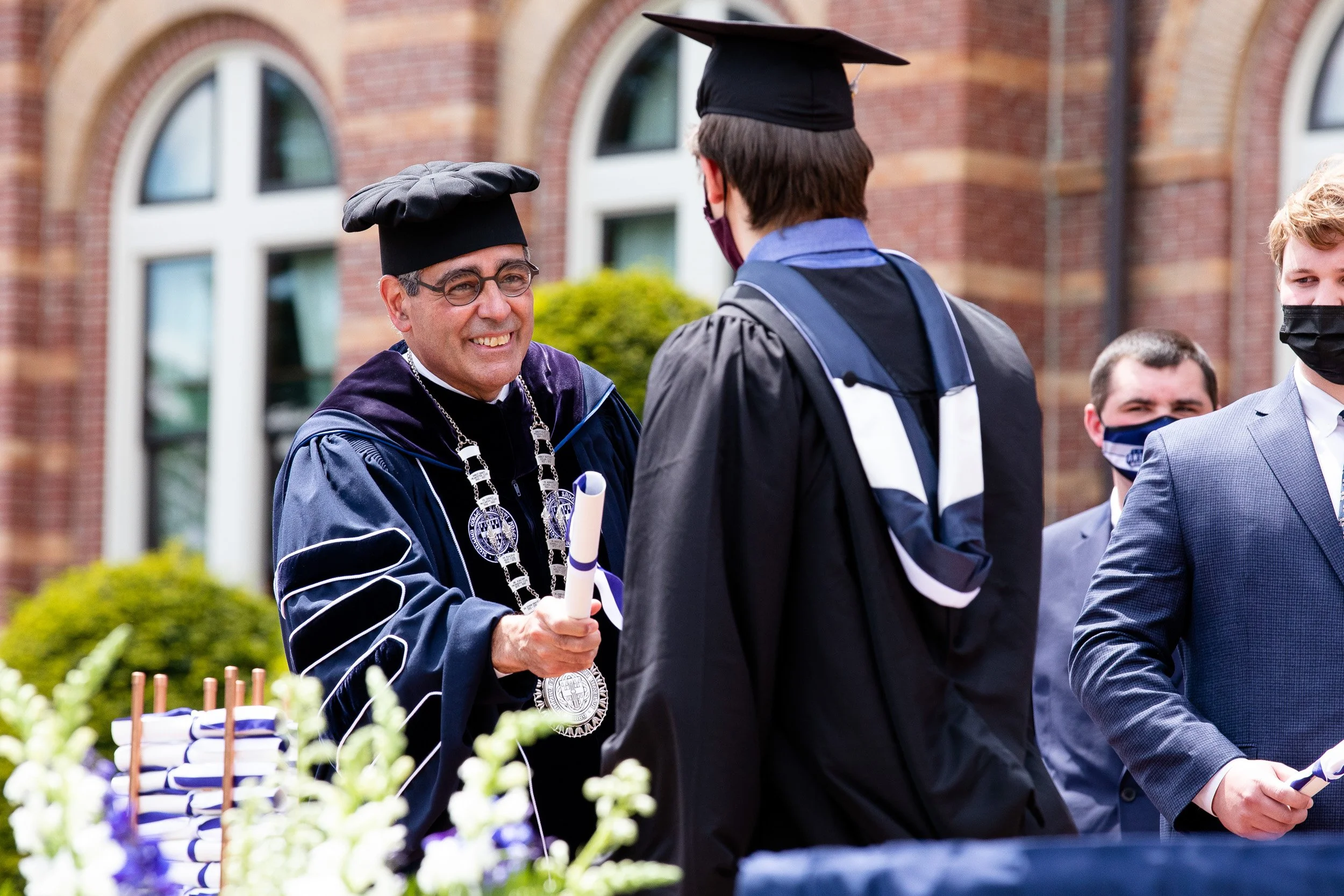 A university graduation ceremony outdoors with faculty in academic regalia handing a diploma to a graduate in cap and gown.