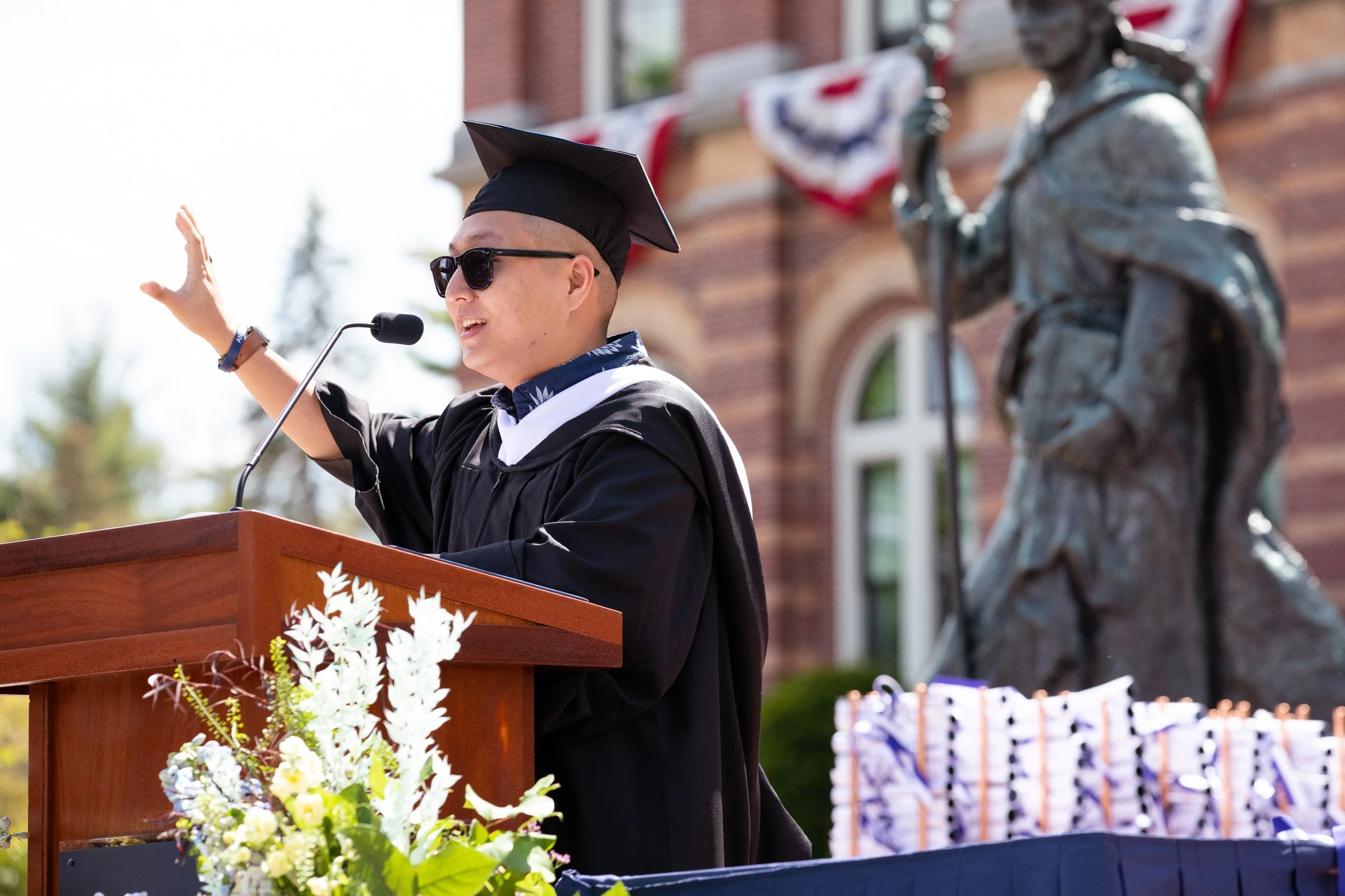 A young man in a cap and sunglasses giving a speech at a graduation ceremony at a New Hampshire College.