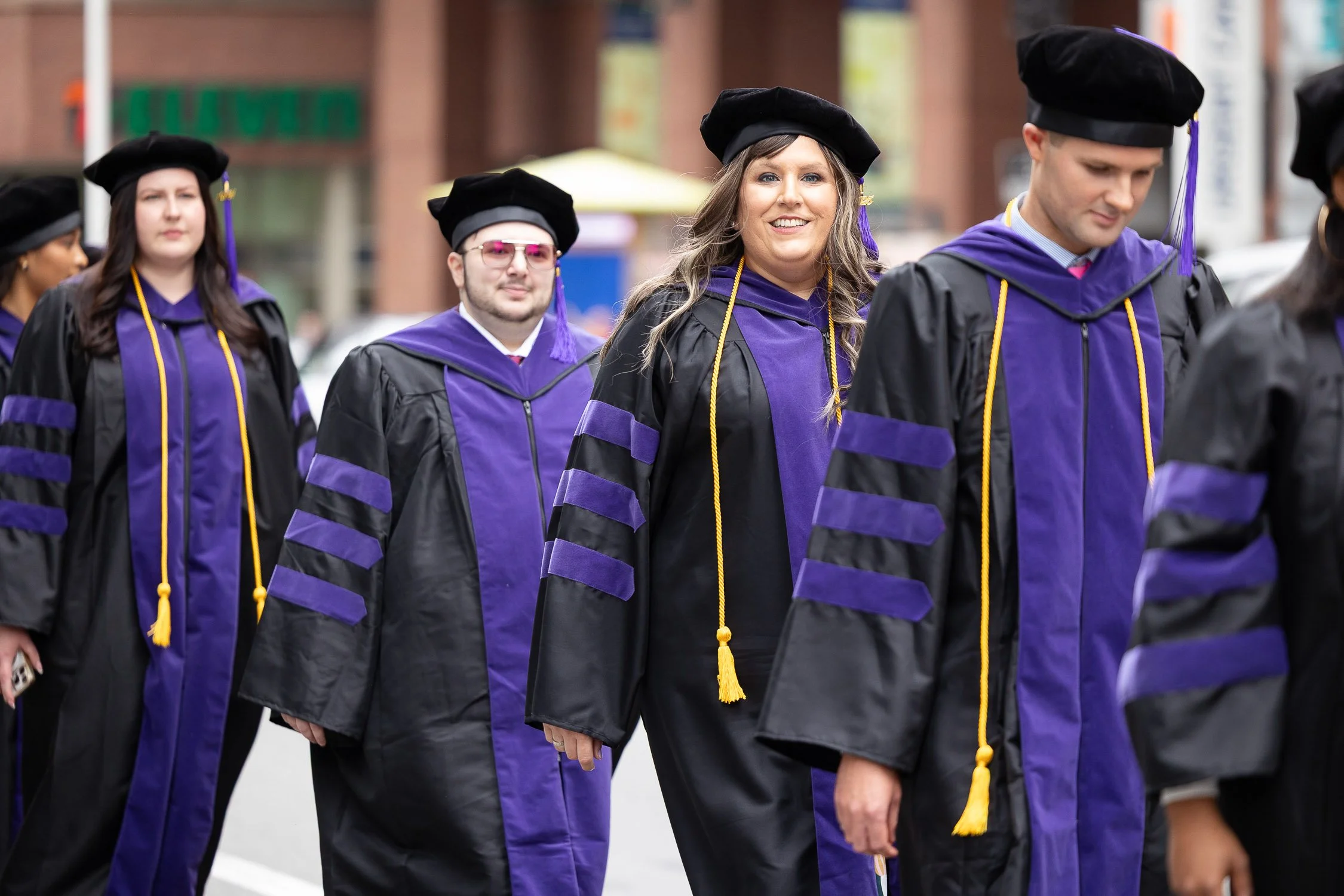 Group of graduates in black robes with purple accents walking in a procession through Boston.