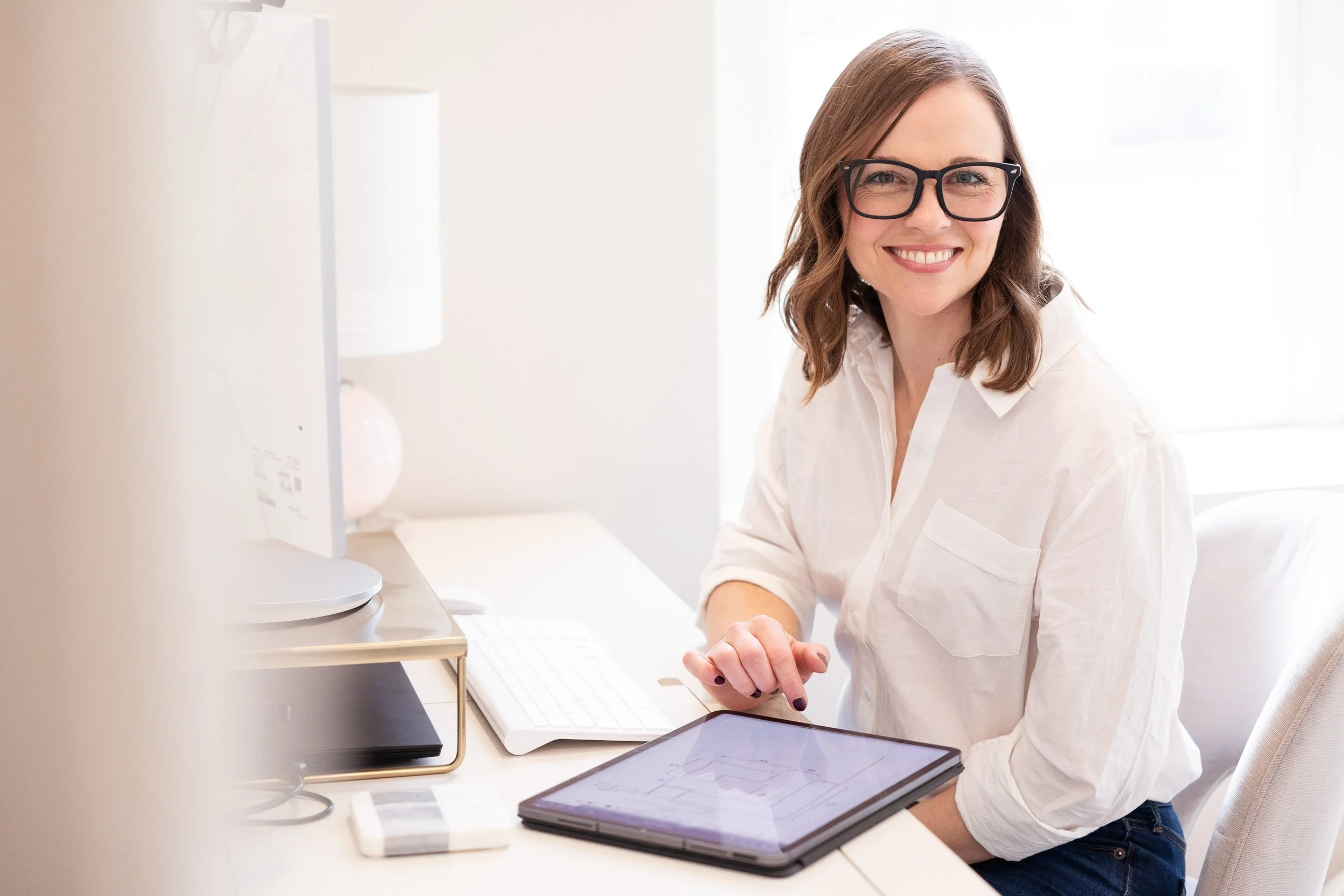 A smiling business woman with glasses and shoulder-length brown hair sitting at a white desk in a bright room, using a digital tablet.