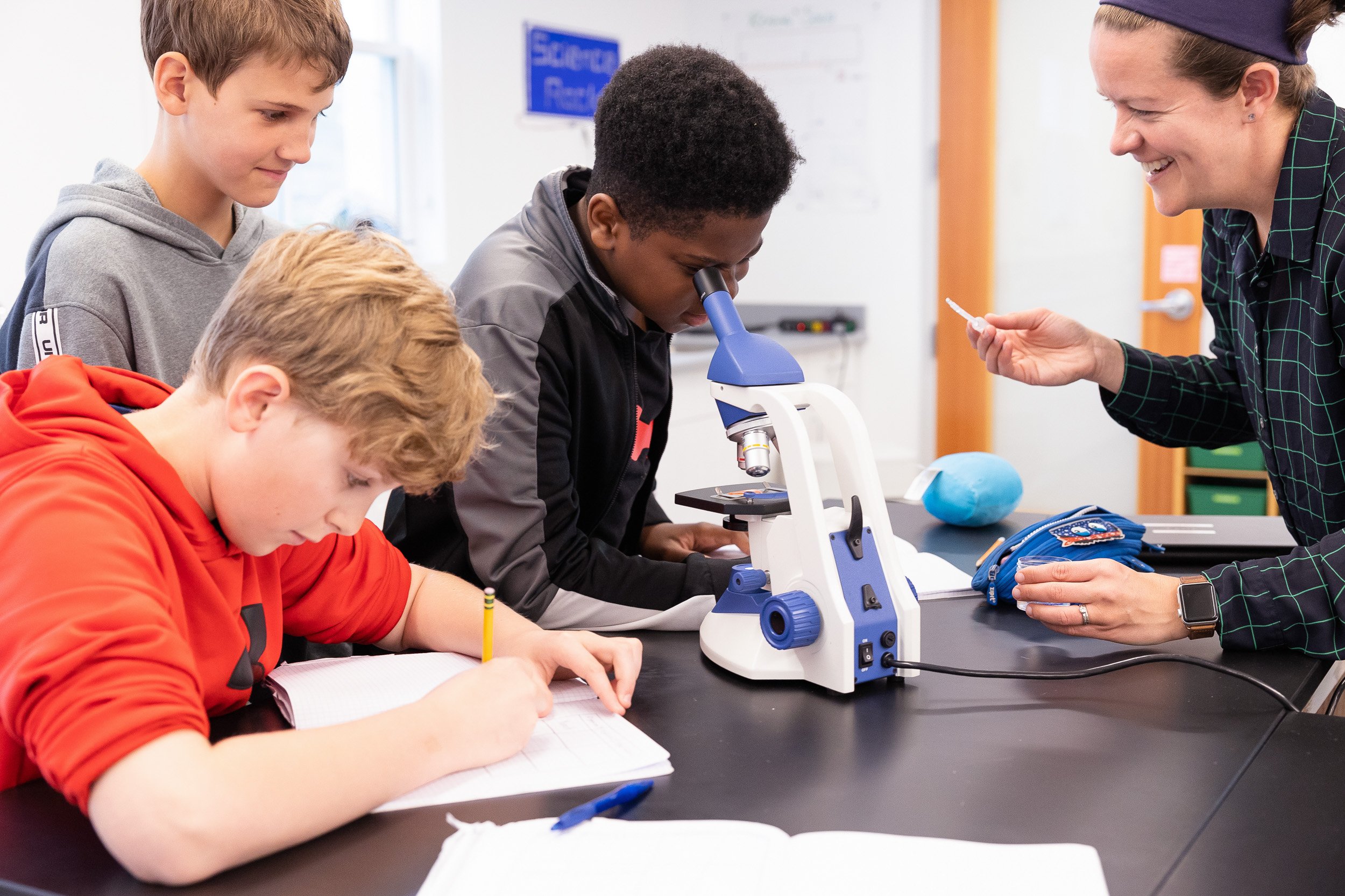 A group of middle school students in a classroom, using a microscope while an instructor engages with the students.