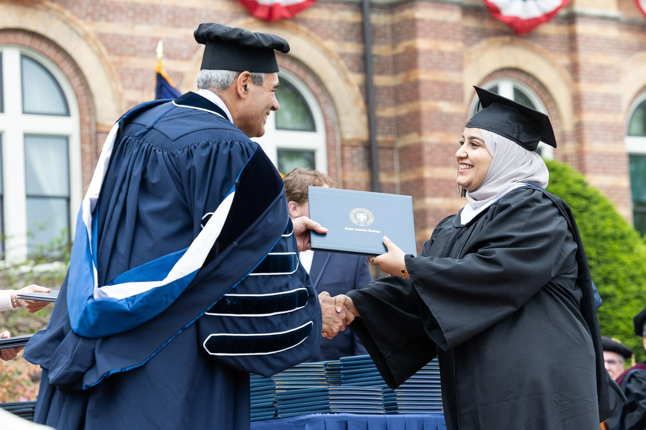 A young woman in a graduation cap and gown receiving her diploma during a graduation ceremony outdoors at a college campus.