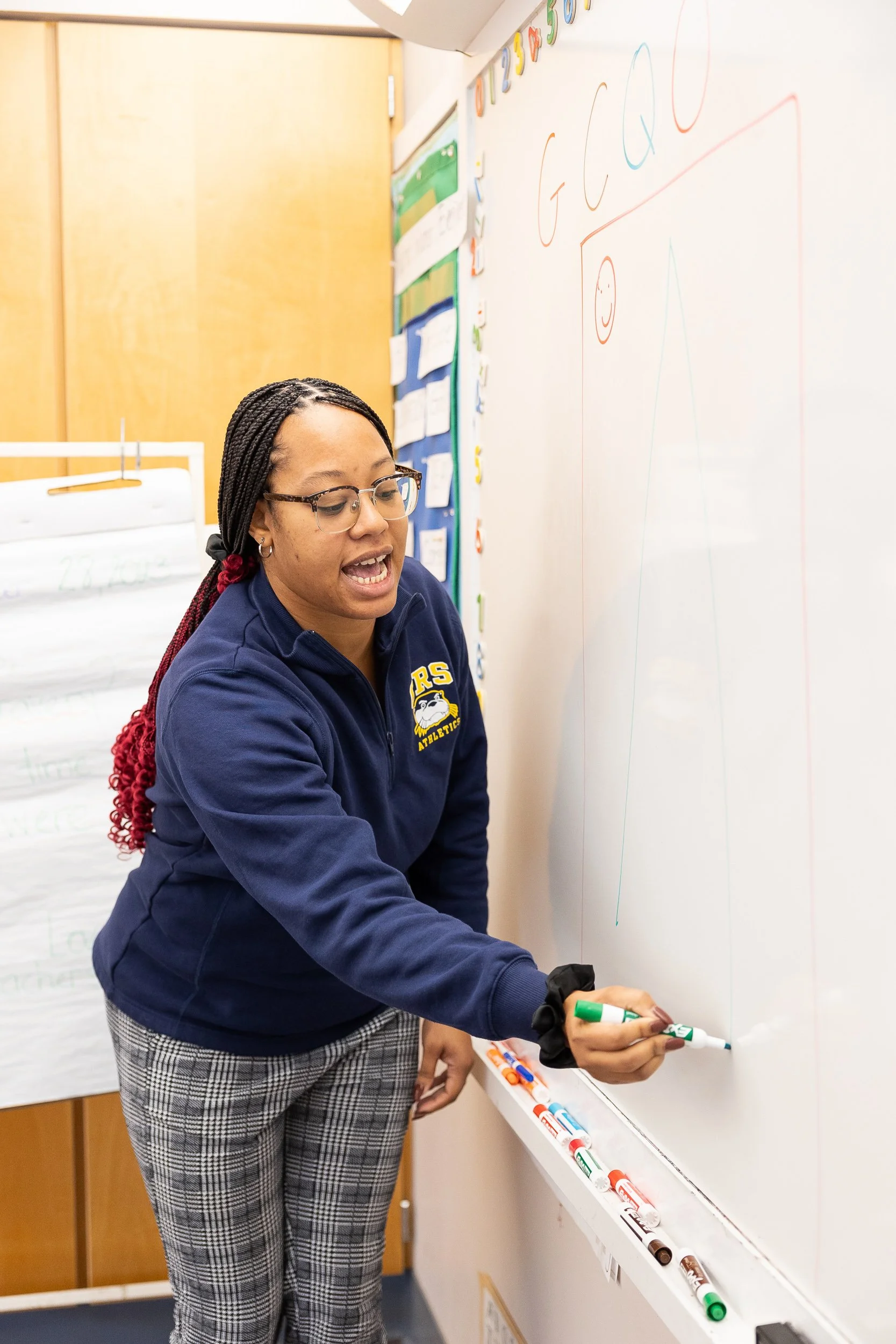 A kindergarten teacher writes on a whiteboard with colored markers in a classroom.