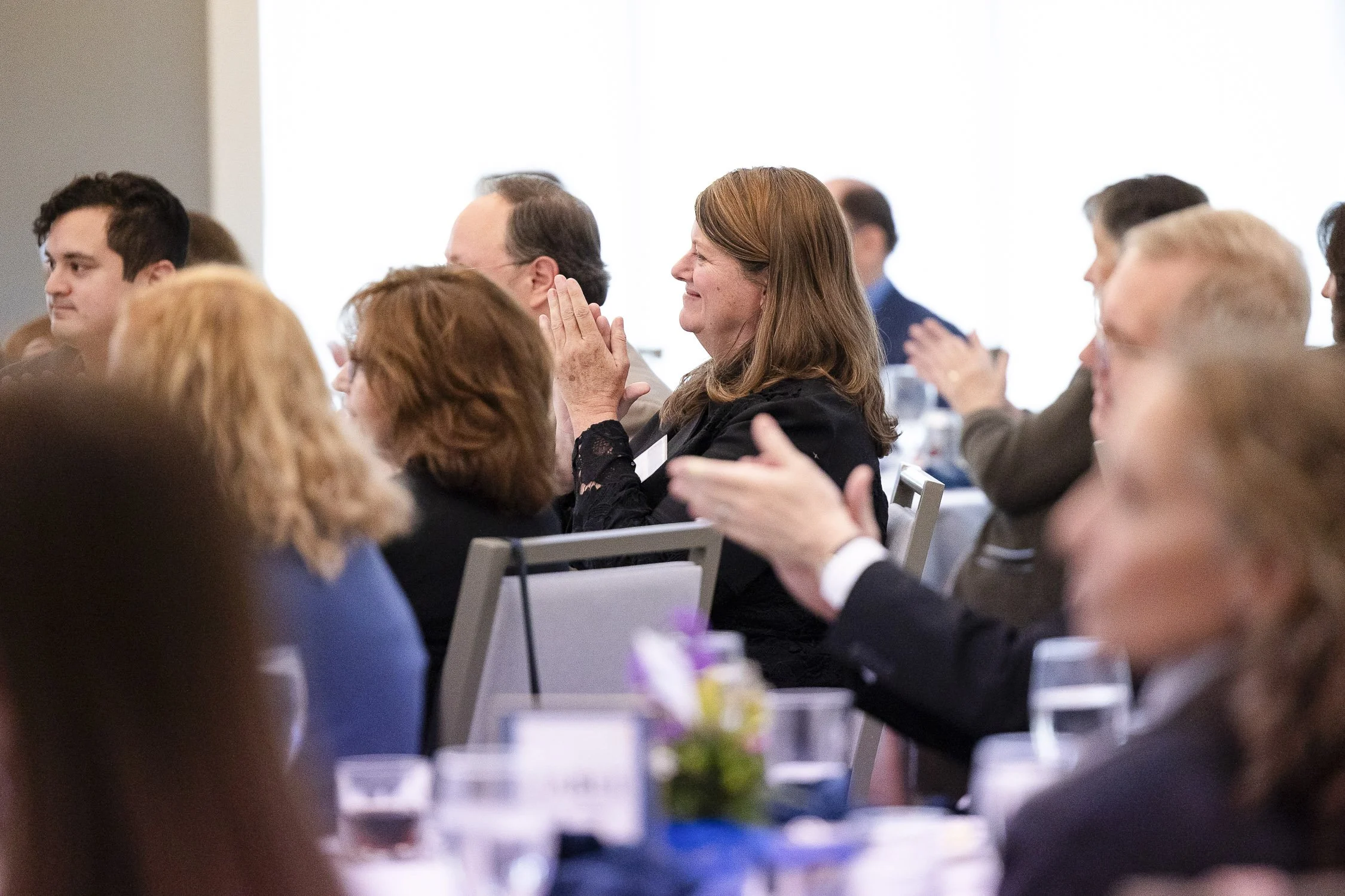 People attending a formal event, sitting at tables, applauding and smiling.