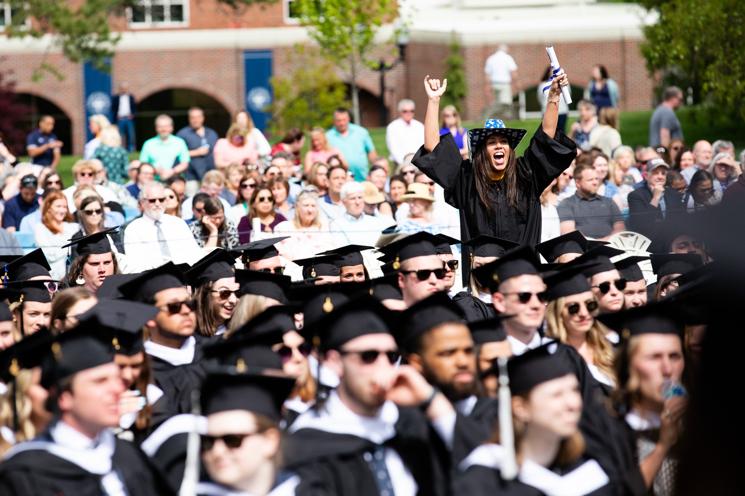 Woman in cap and gown celebrating with her arms raised at outdoor graduation ceremony at Saint Anselm College.