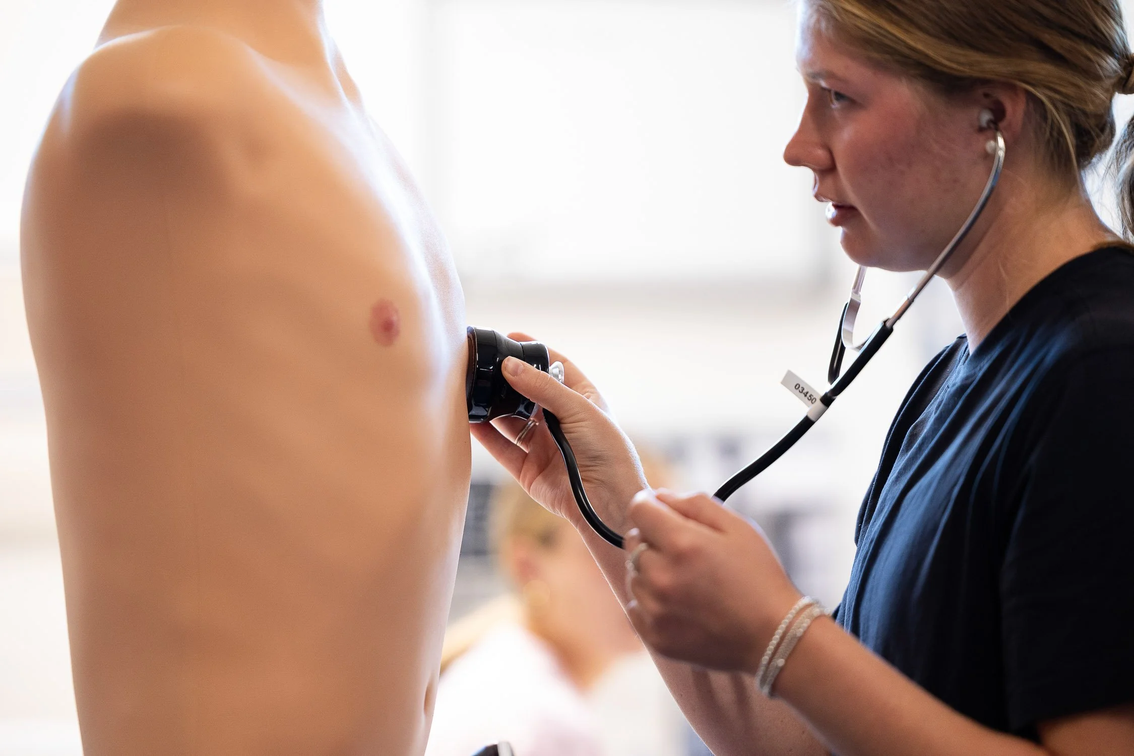 A college nursing student using a stethoscope on a medical training mannequin.