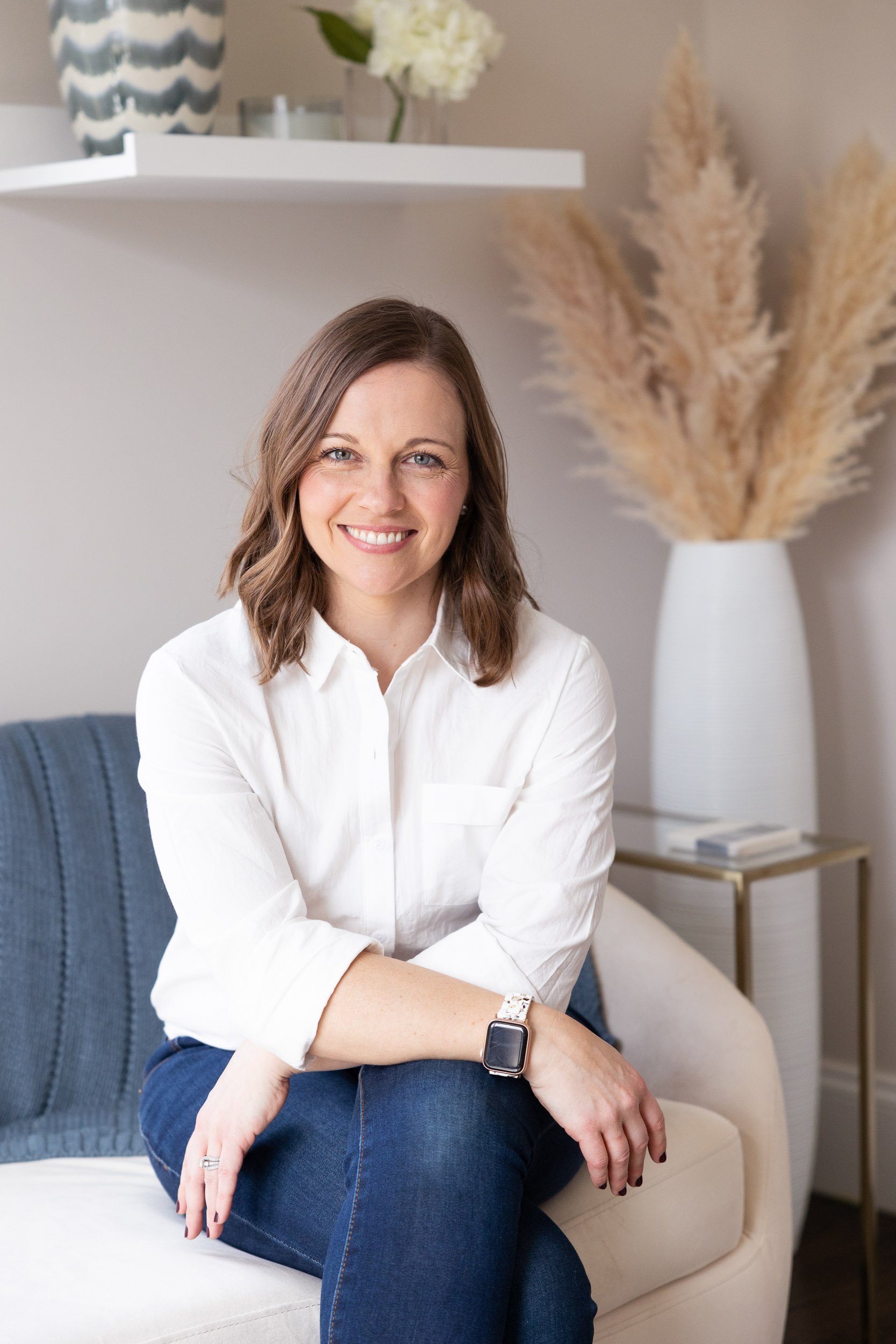 A Massachusetts interior designer with shoulder-length brown hair smiling sitting on a beige sofa in a modern living room, wearing a white shirt and blue jeans.