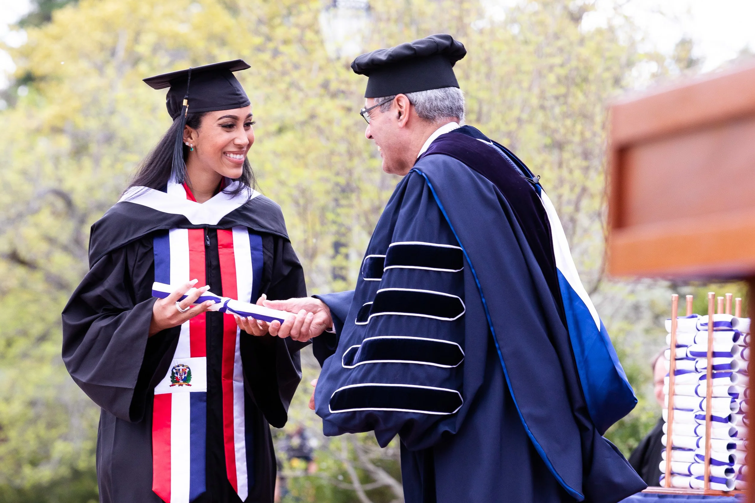 A woman in a graduation gown and cap is receiving her diploma from the college president in an outdoor graduation ceremony.