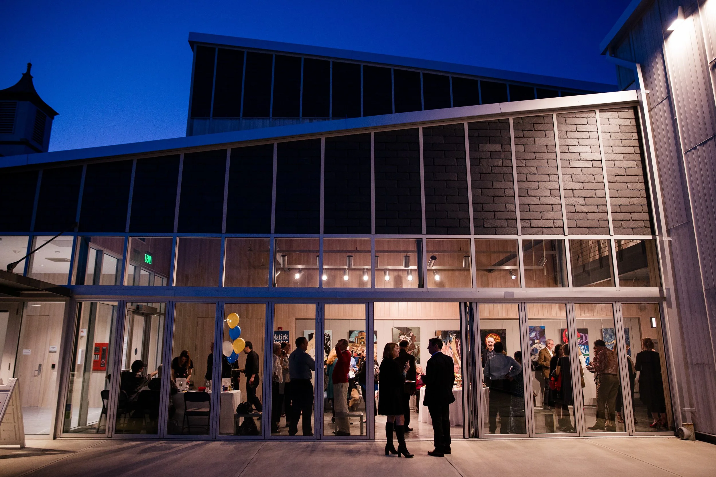 People gathered inside a modern building with glass walls during an evening event at Walnut Hill School for the Arts in Natick.