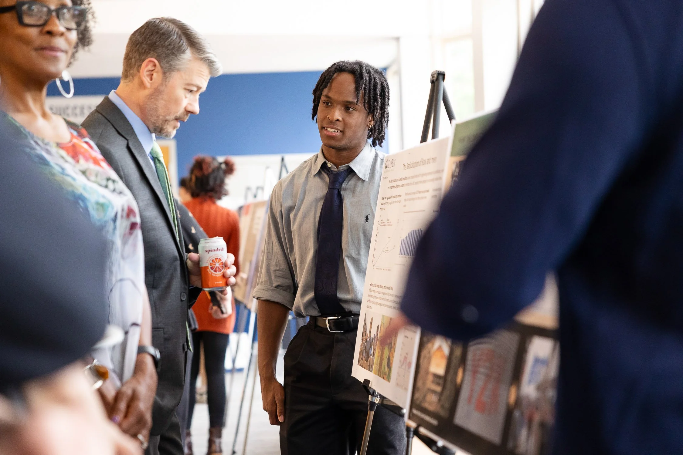 A young man in a collared shirt explains a scientific poster to a group at MassBay Community College.