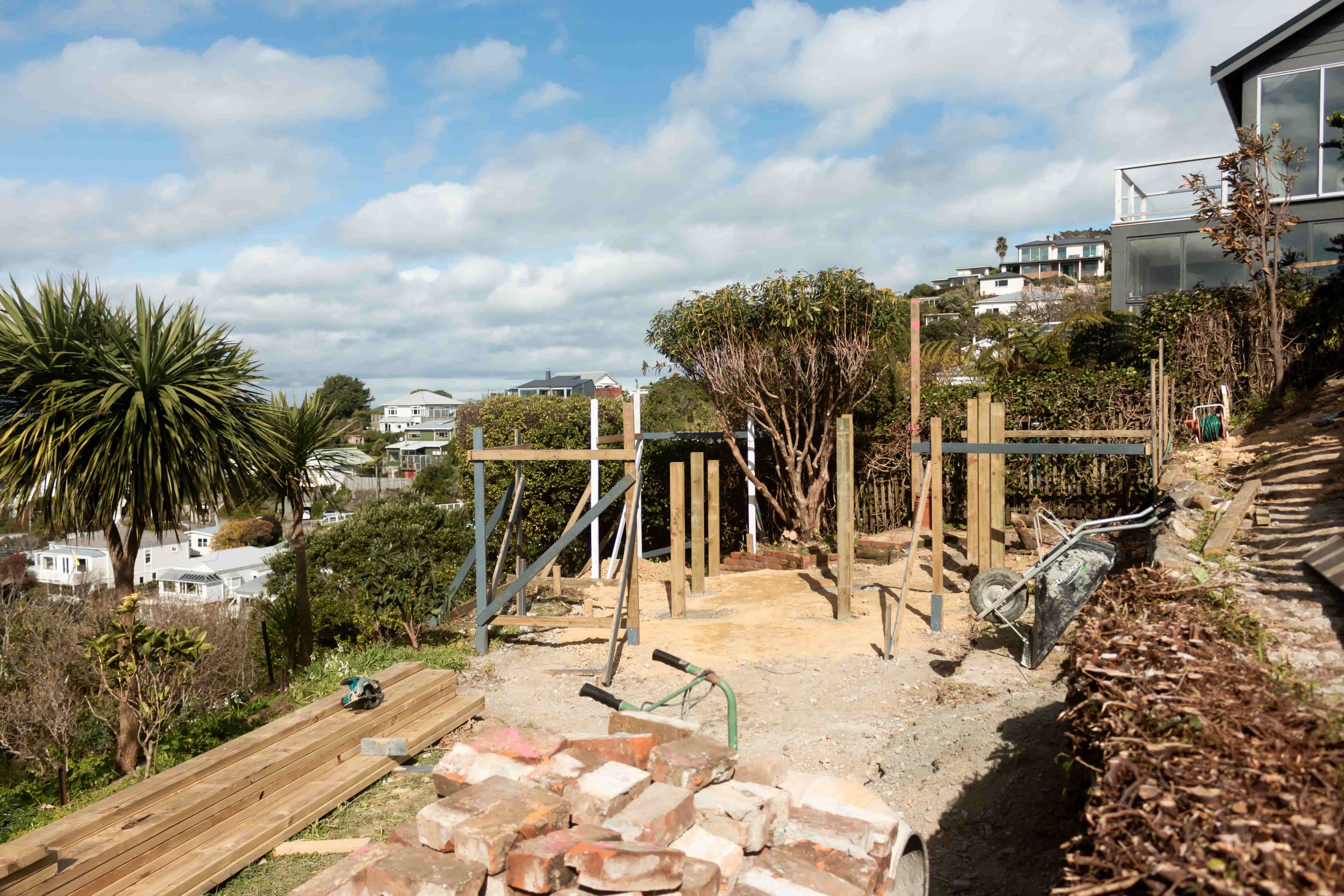 Construction site on a hillside with scattered building materials, wooden posts, metal framing, and a wheelbarrow, residential houses in the background under a partly cloudy sky.