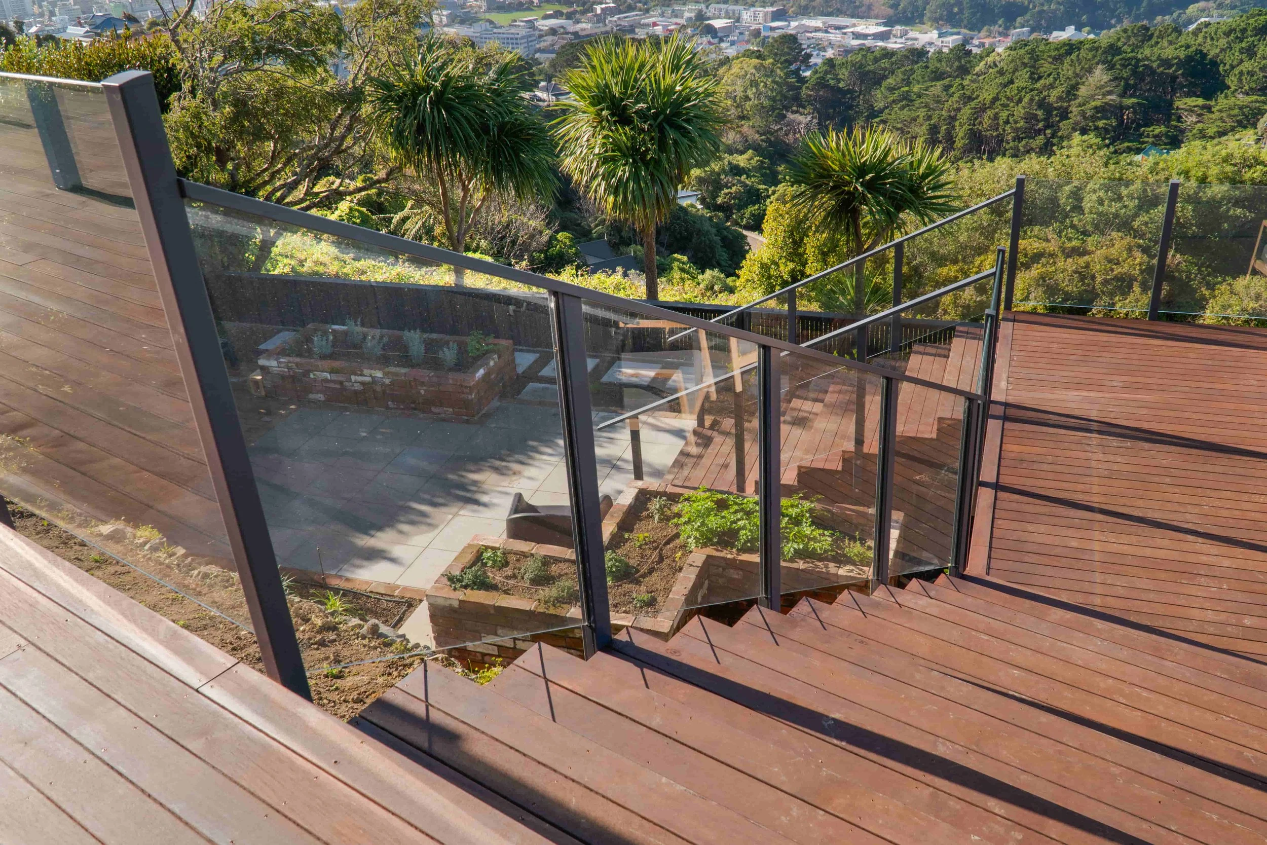 A view of a wooden deck with glass railing overlooking a garden with trees and potted plants, and a distant view of a city in the background.