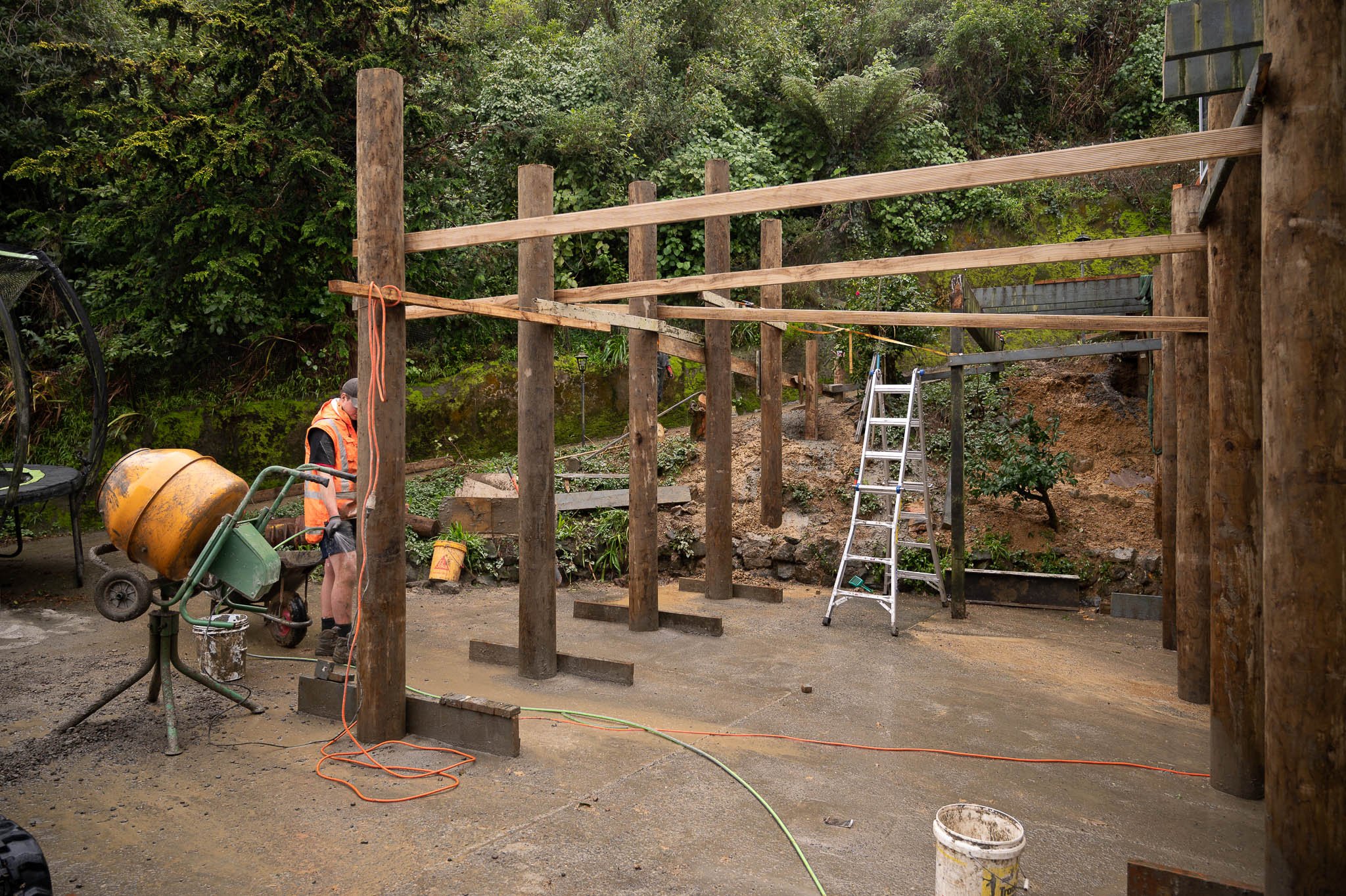 Construction site with wooden posts and beams, a worker in safety vest next to concrete mixer, ladder, and construction tools, surrounded by dirt and greenery.