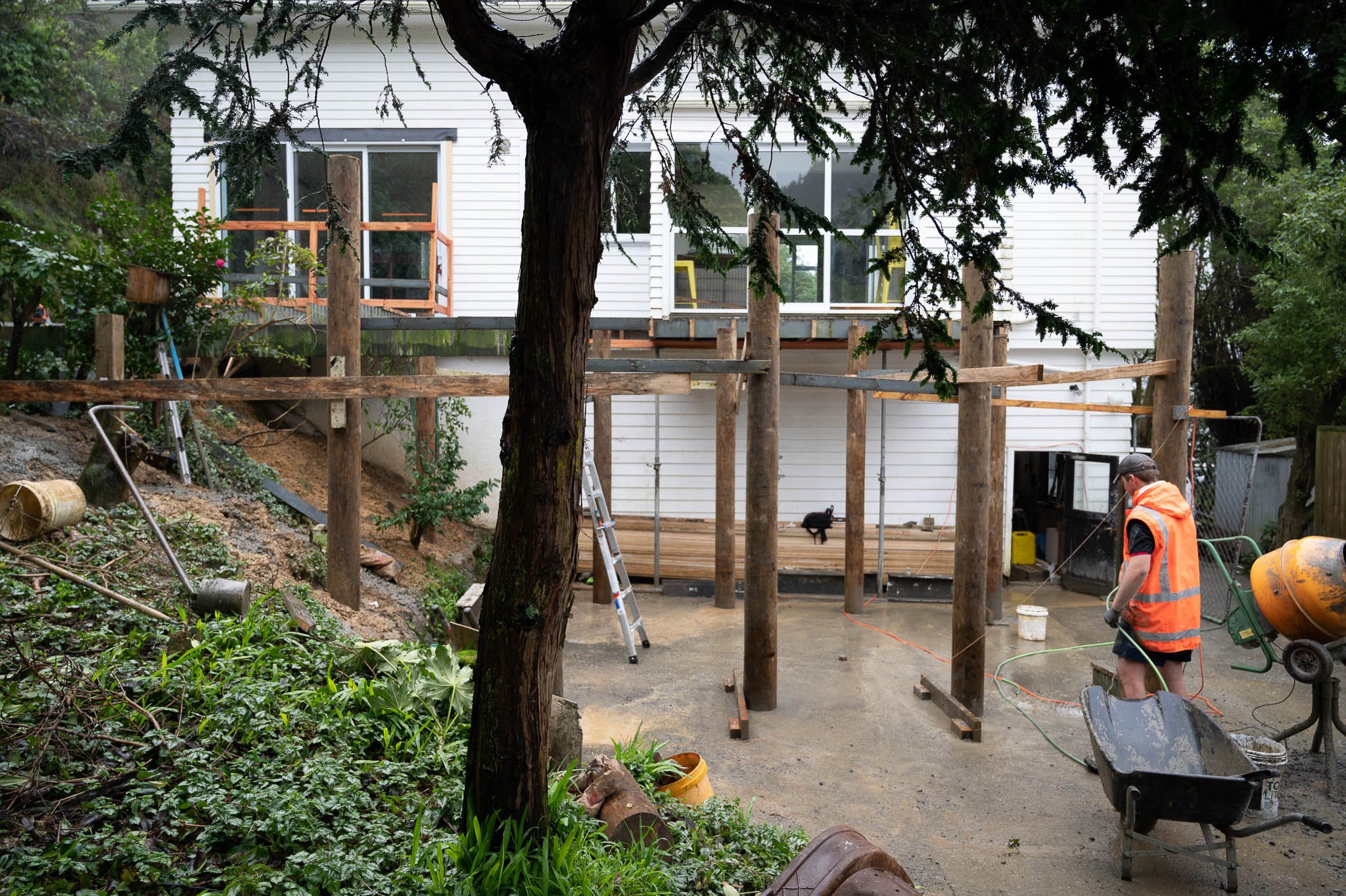 Construction worker in an orange safety vest working on building a deck or patio area outside a white house, with construction materials and tools around, and trees and greenery in the background.