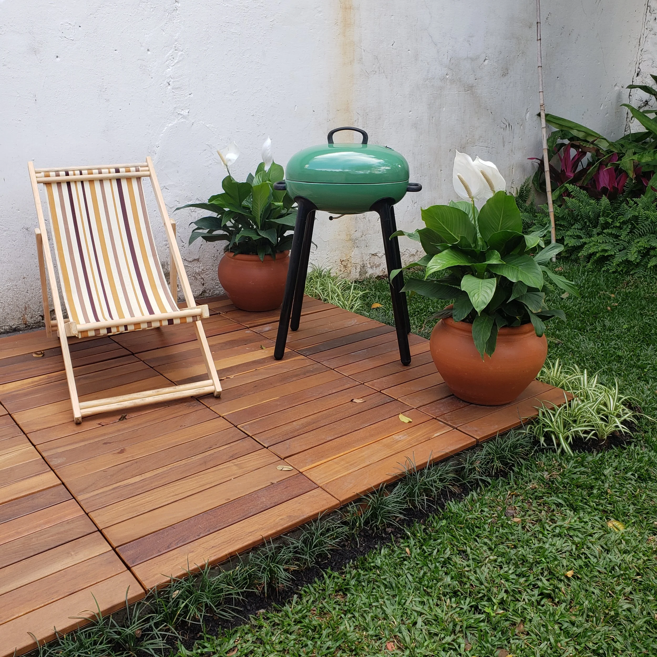 Área de varanda com piso de madeira, duas plantas em vasos de barro com flores brancas, uma churrasqueira portátil verde, uma cadeira de praia de madeira com encosto de tecido listrado e cerca de grama verde.