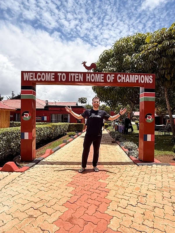 Homme souriant avec bras ouverts devant une entrée en arc rouge marquée "Welcome to Iten Home of Champions", avec des arbres et des bâtiments en arrière-plan.