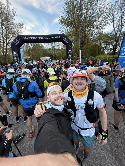 Louis Gronemberg et un ami, souriants qui prennent un selfie avant une course, entourés de coureurs, avec une arche de départ noire portant l'inscription 'SkyRace des Matheysins'.