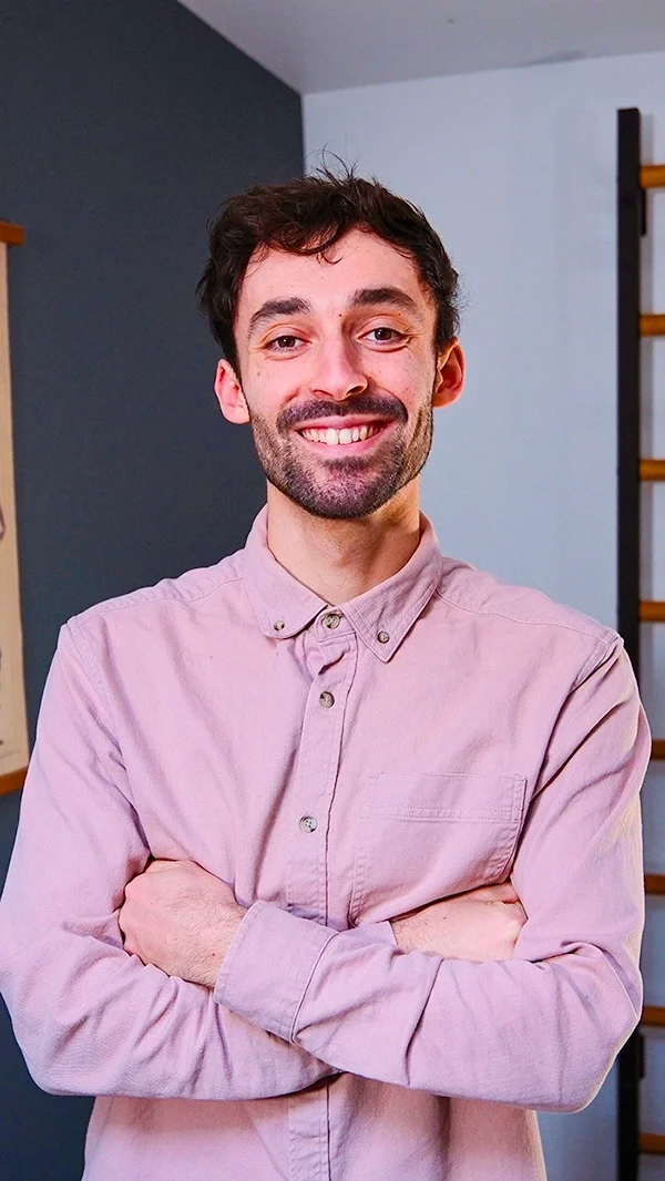 Nicolas Ménard, jeune homme souriant avec bras croisés, portant une chemise rose clair, dans un intérieur moderne.